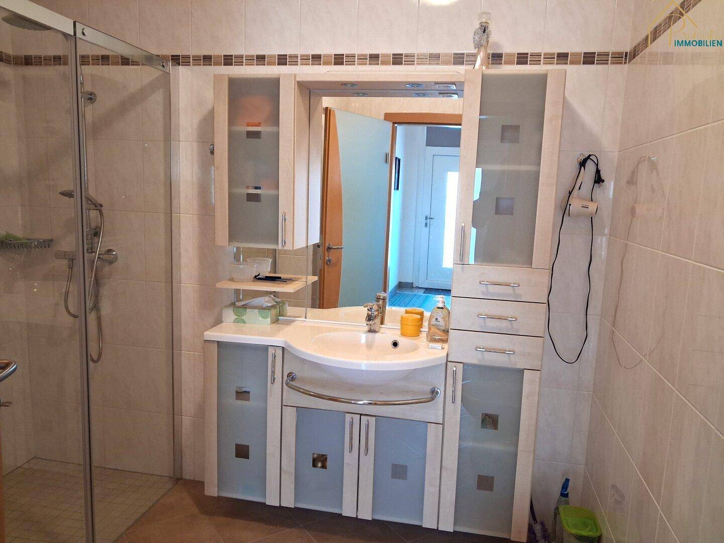 Badezimmer mit Waschtischunterschrank, Spiegel und Duschkabine in hellen Fliesen.