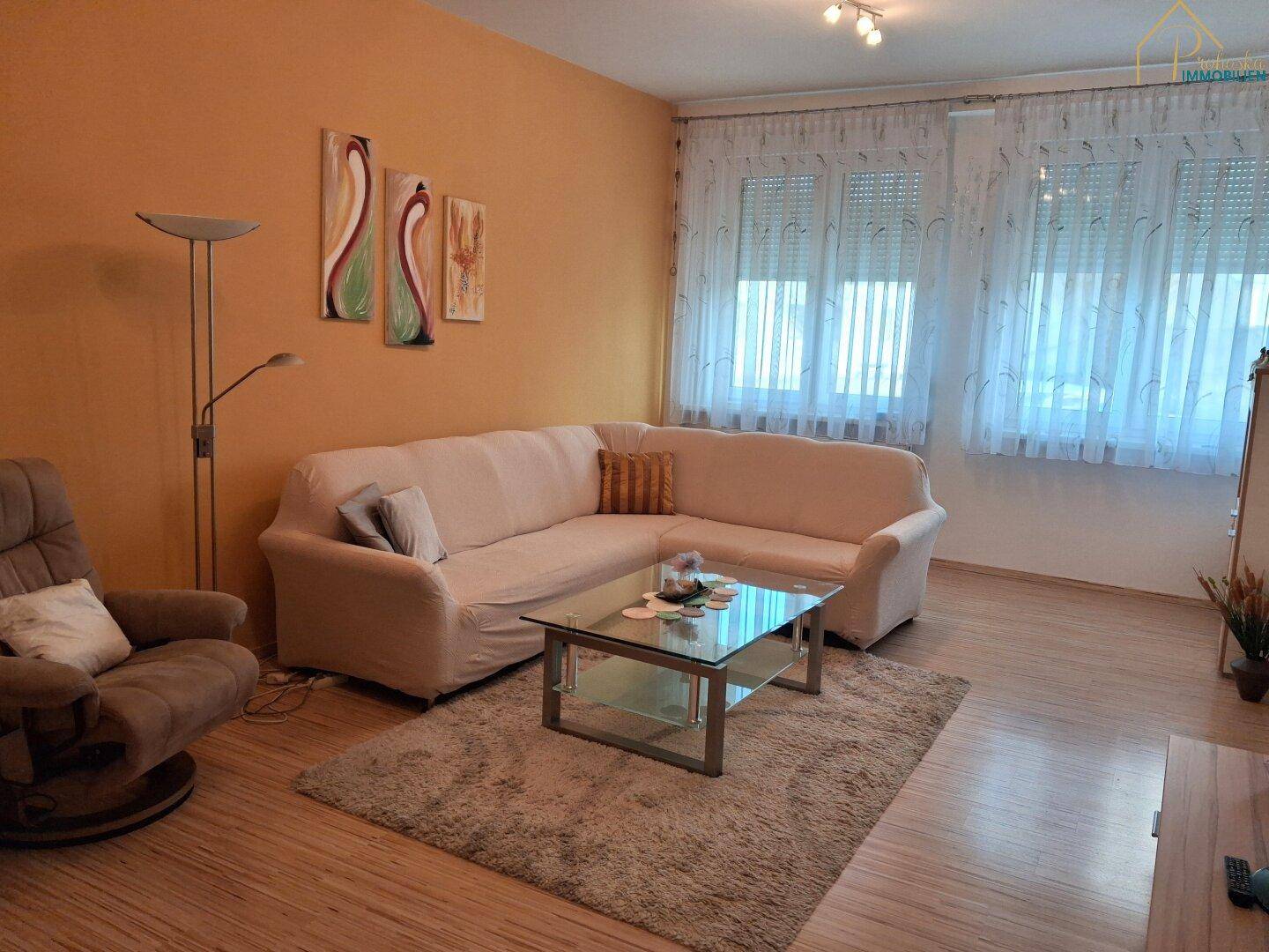 Geräumiges Wohnzimmer mit beigem Ecksofa, Couchtisch und großen Fenstern mit Vorhängen.