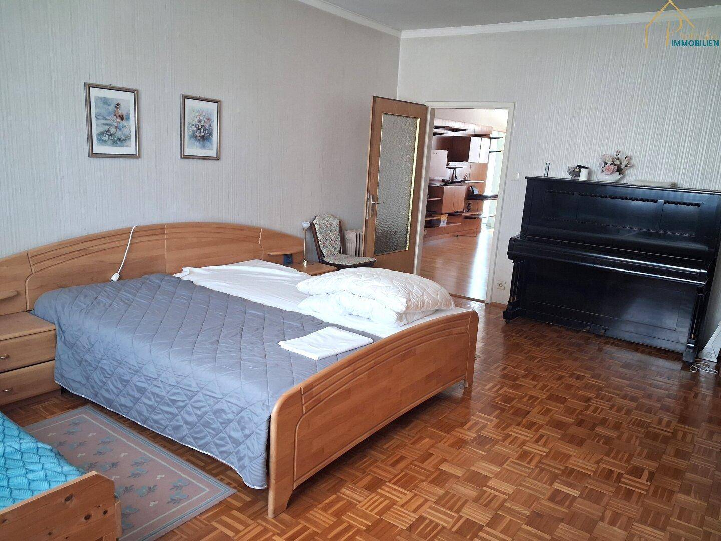Schlafzimmer mit Holzbett, Klavier und Parkettboden, mit Zugang zu einem weiteren Raum.