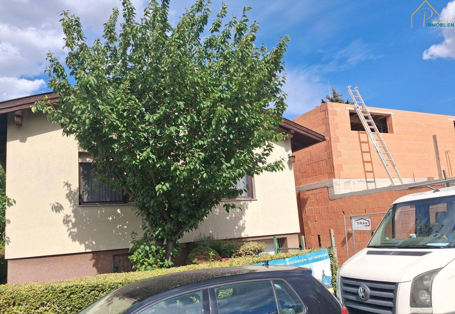 Hausfassade mit Baum und angrenzender Baustelle unter blauem Himmel.