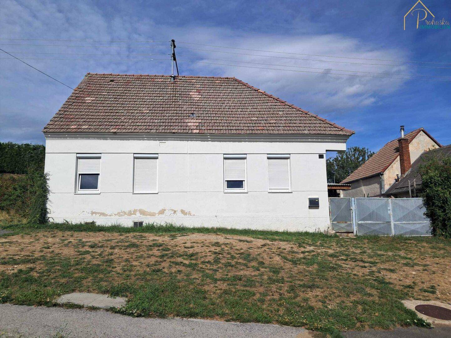 Vorderansicht eines weißen Hauses mit Ziegeldach und Rollläden in ländlicher Umgebung.