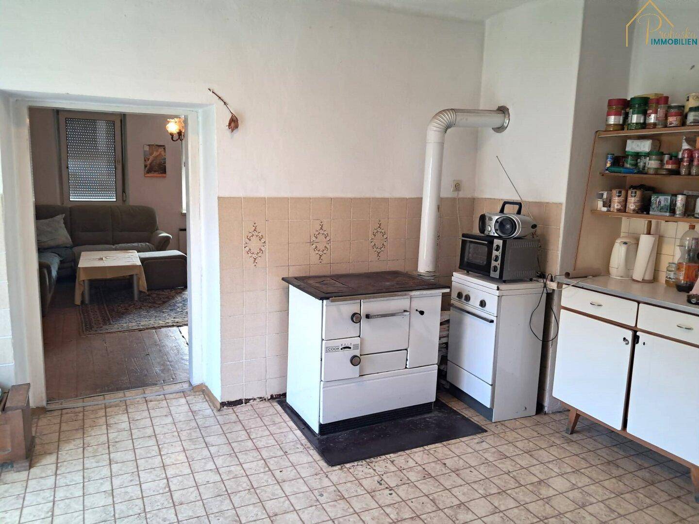 Rustikale Küche mit Fliesenboden, Holzofen und Blick in das angrenzende Wohnzimmer.
