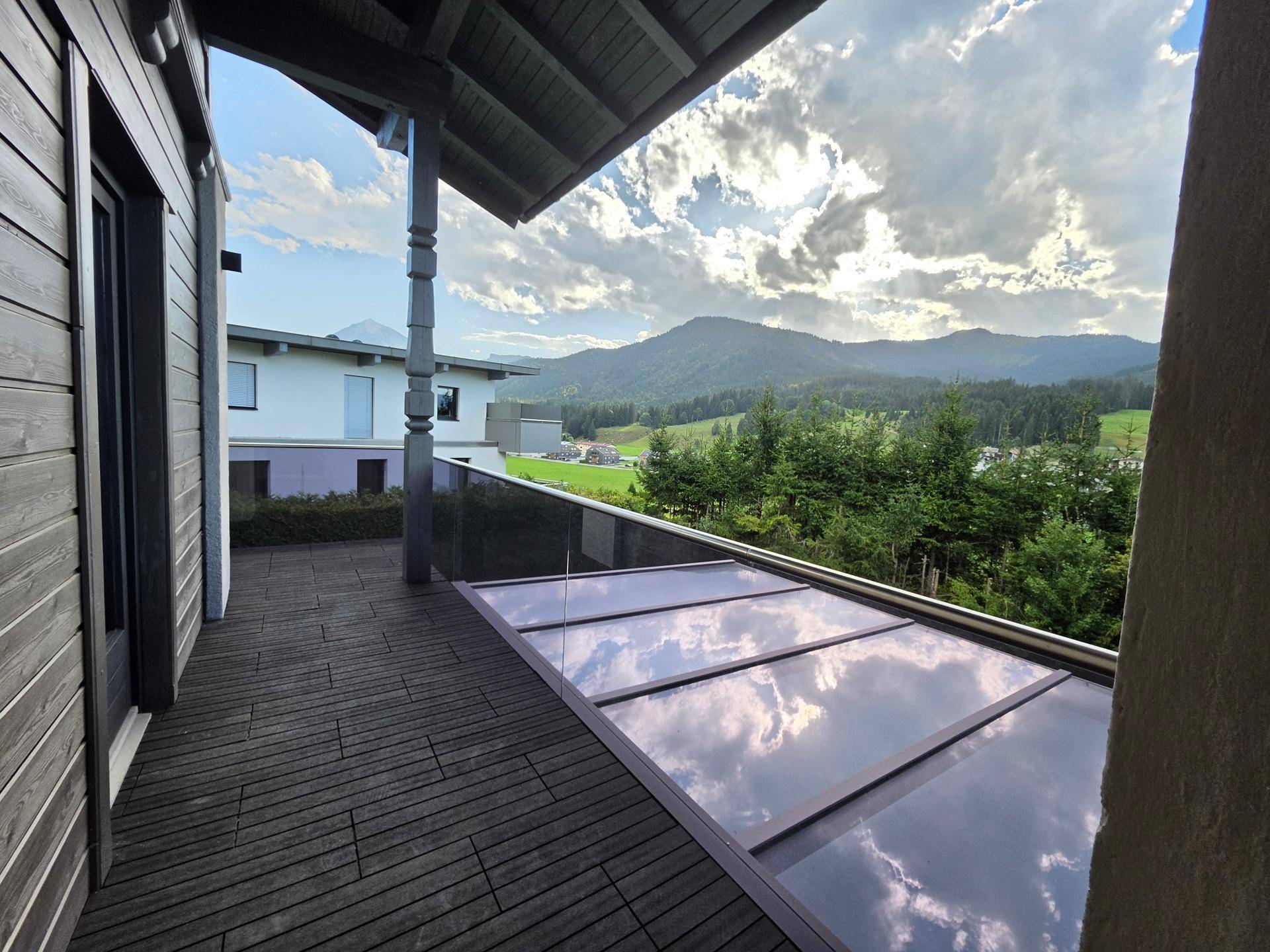 Geräumiger Balkon mit Holzboden, Glasgeländer und schützendem Glasdach mit Bergblick.