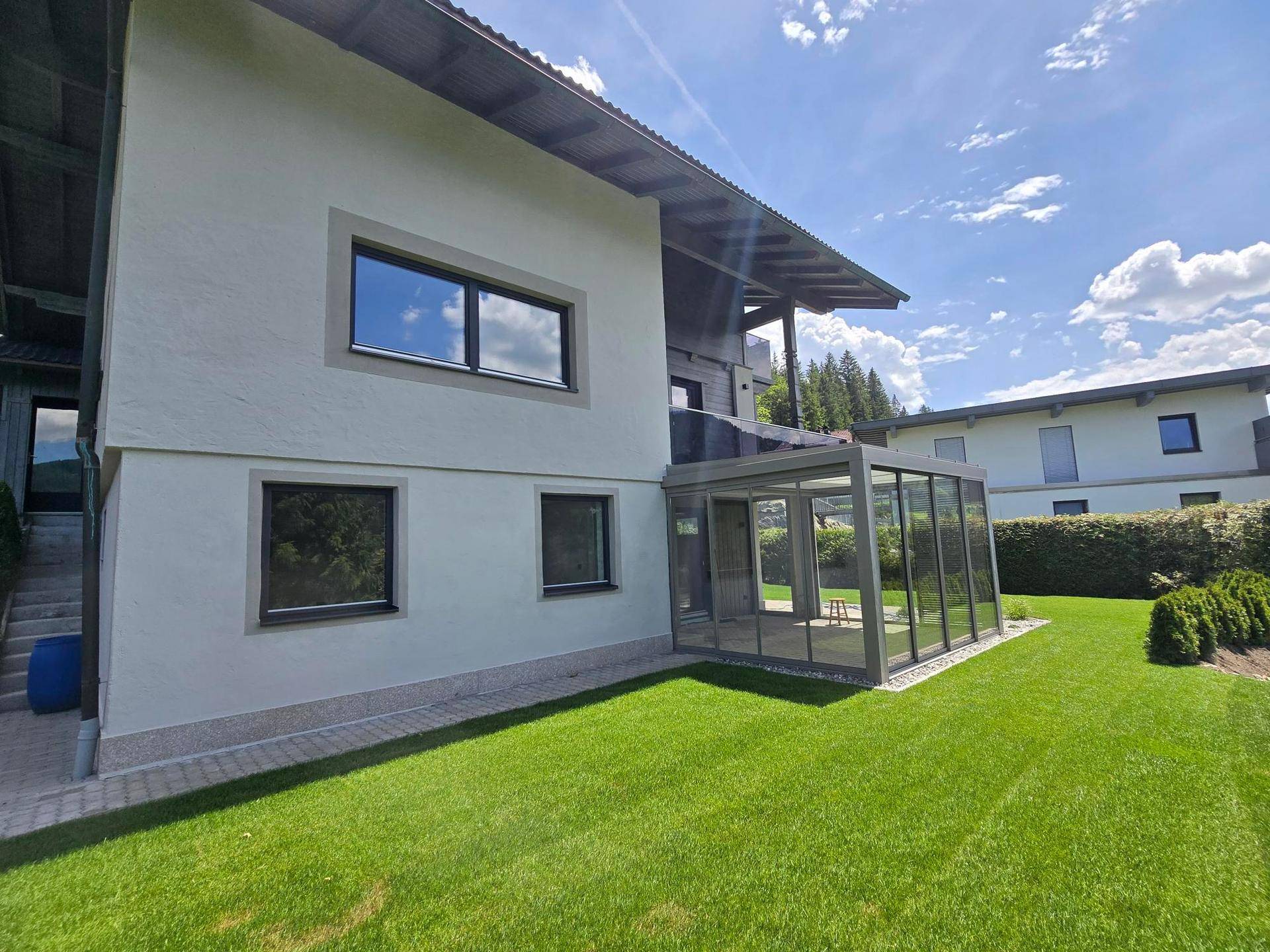Rückansicht des Hauses mit Glasanbau und weitläufigem Gartenbereich unter blauem Himmel.