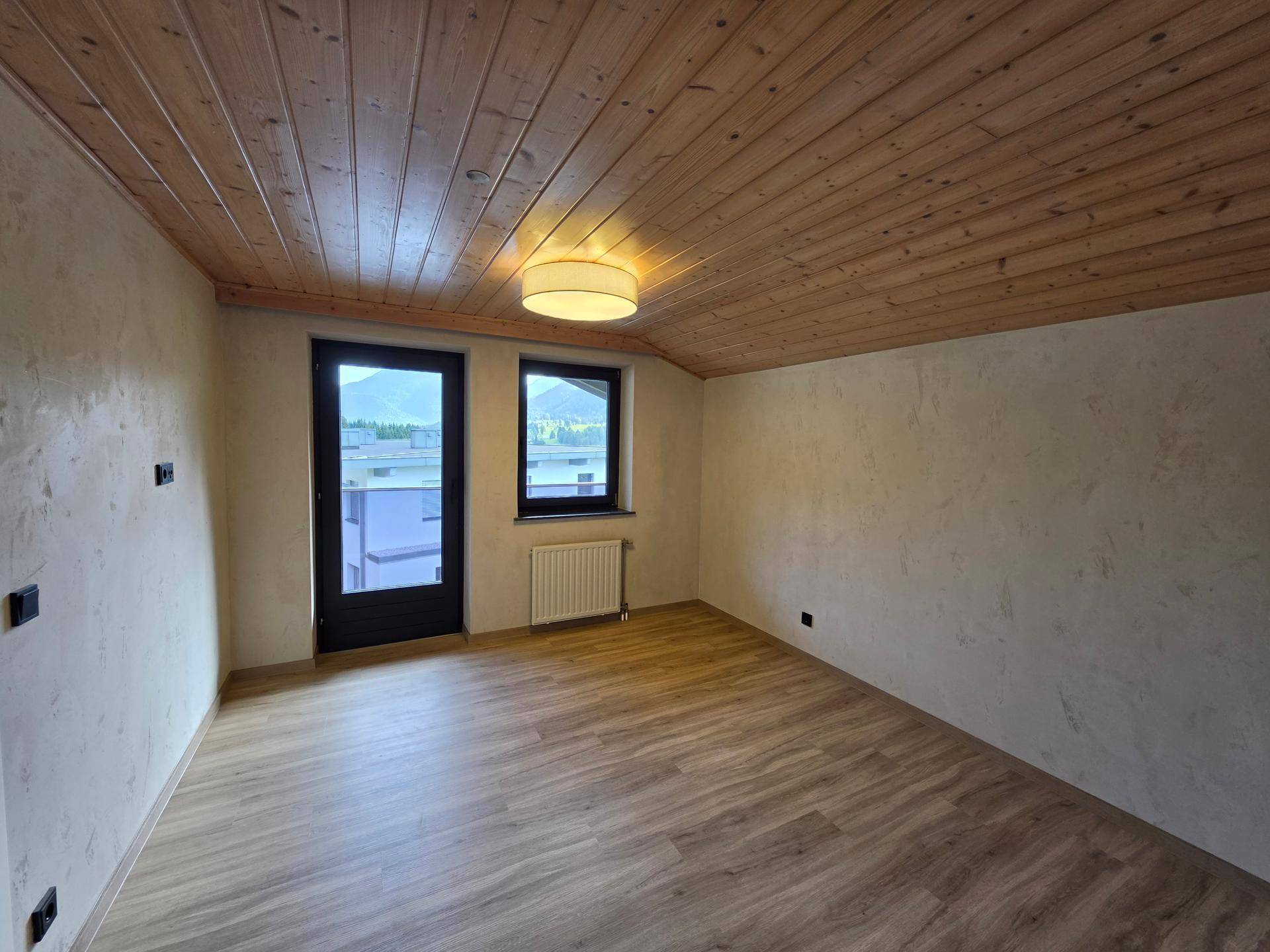Helles Dachgeschosszimmer mit Holzdecke, Laminatboden und Zugang zu einem Balkon.