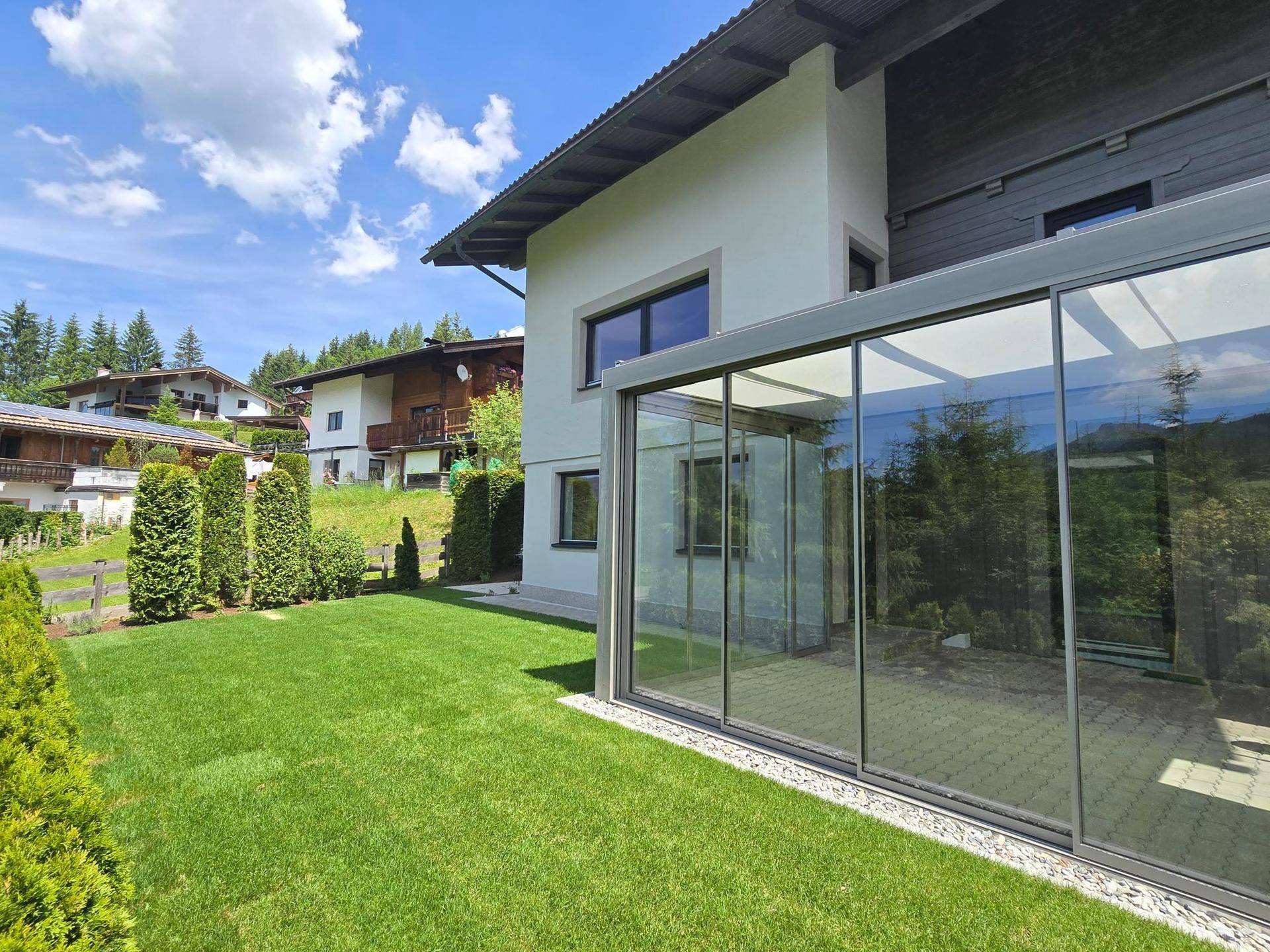 Hausfassade mit modernem Glasanbau und weitläufiger, grüner Rasenfläche.