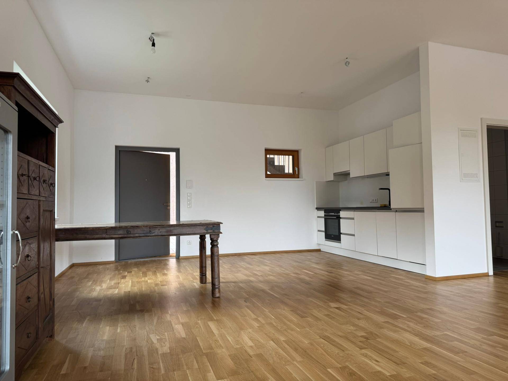 Offener Wohnbereich mit integrierter, moderner weißer Küche und Holzboden, ideal für geselliges Beisammensein.