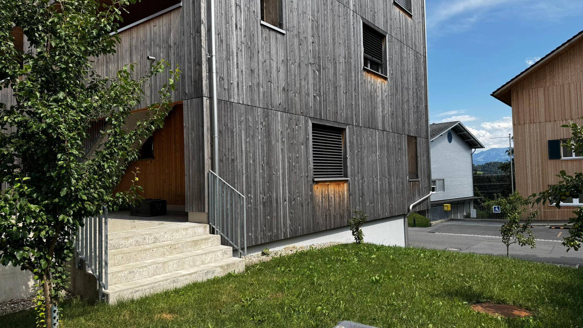 Gebäudeeingang mit Holzfassade und Steintreppe, umgeben von Grünflächen unter blauem Himmel.
