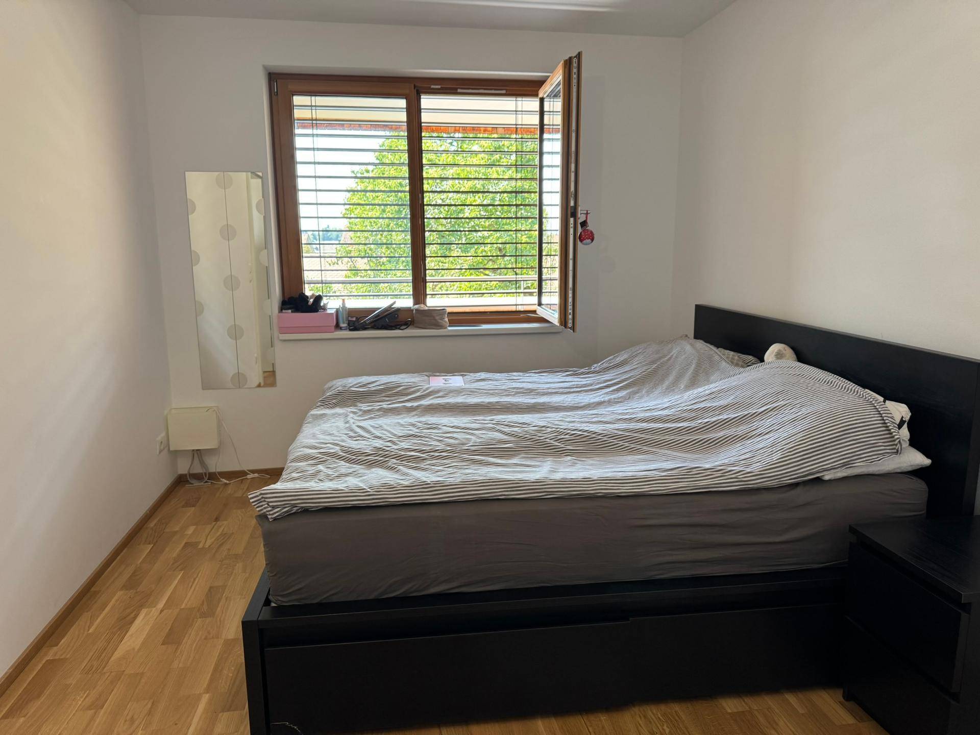 Schlafzimmer mit Holzboden, Doppelbett und Fenster mit Jalousien und Blick ins Grüne.