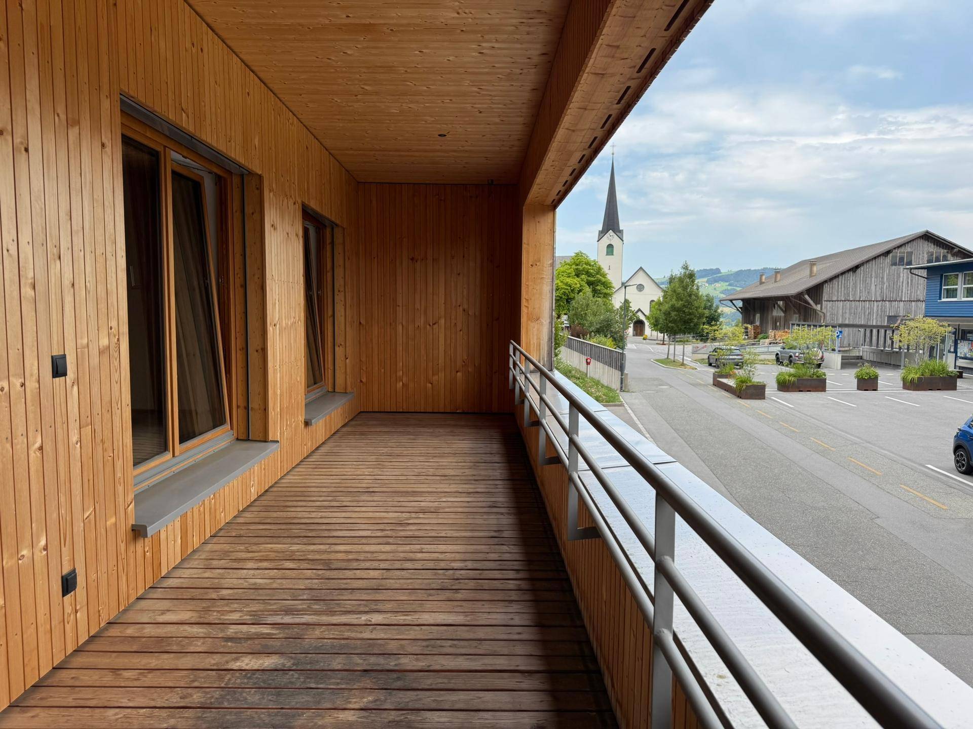 Geräumiger Balkon mit Holzboden und Blick auf die Straße und die umliegende Bebauung mit Kirchturm.