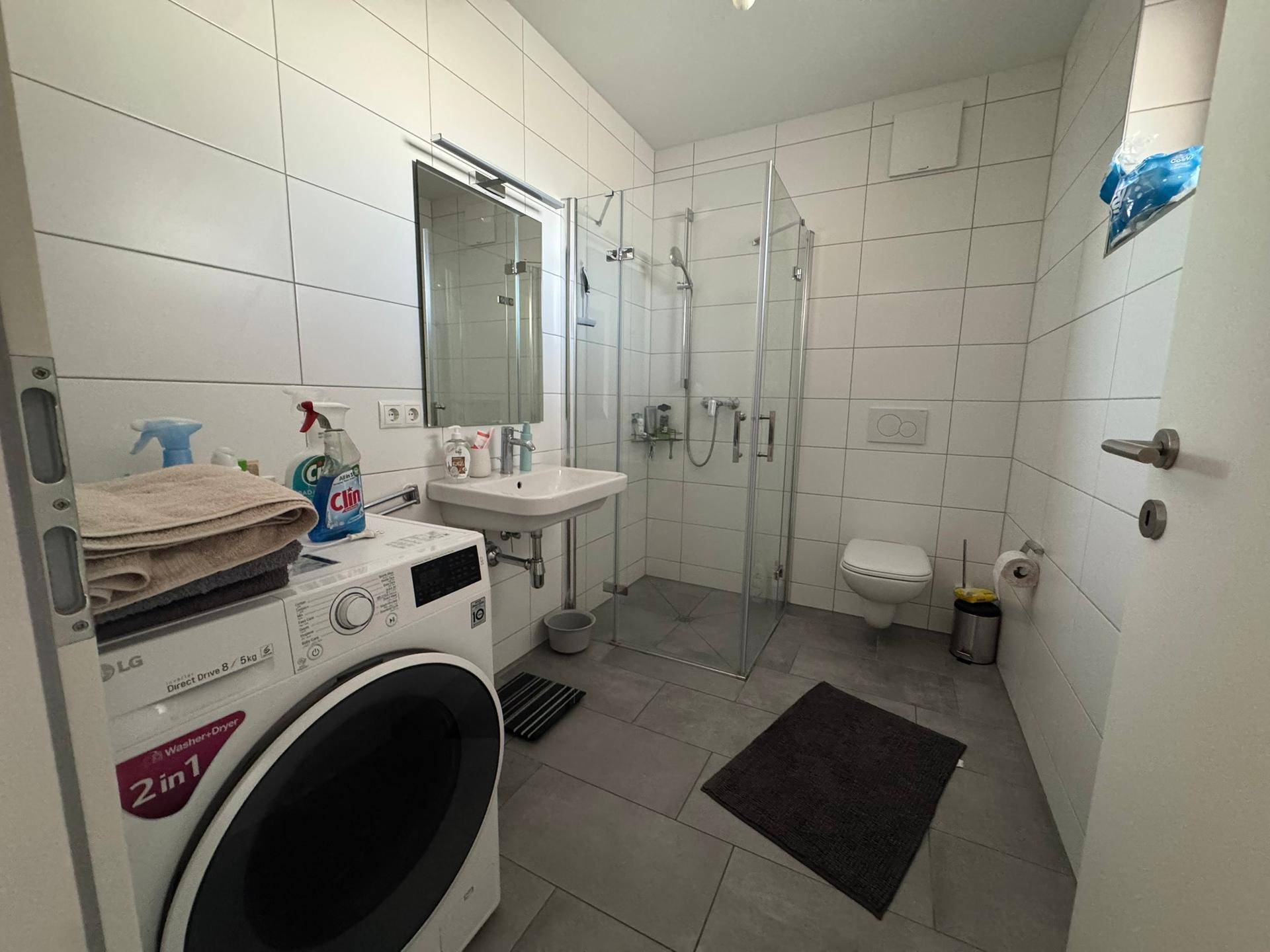 Modernes Badezimmer mit weißen Fliesen, Duschkabine, Waschbecken und integrierter Waschmaschine.