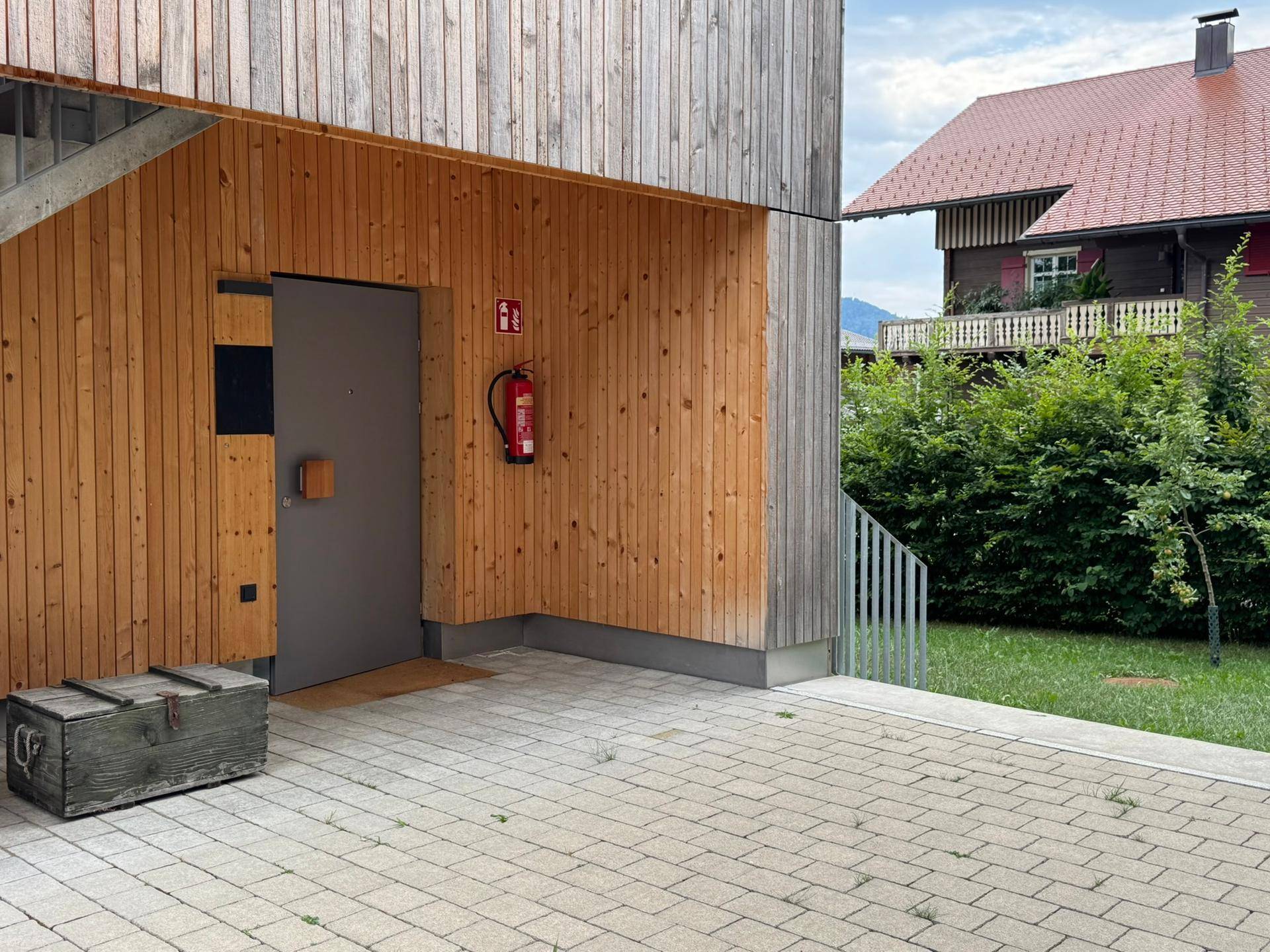 Eingangsbereich mit Holzverkleidung und gepflastertem Vorplatz, ausgestattet mit einem Feuerlöscher.