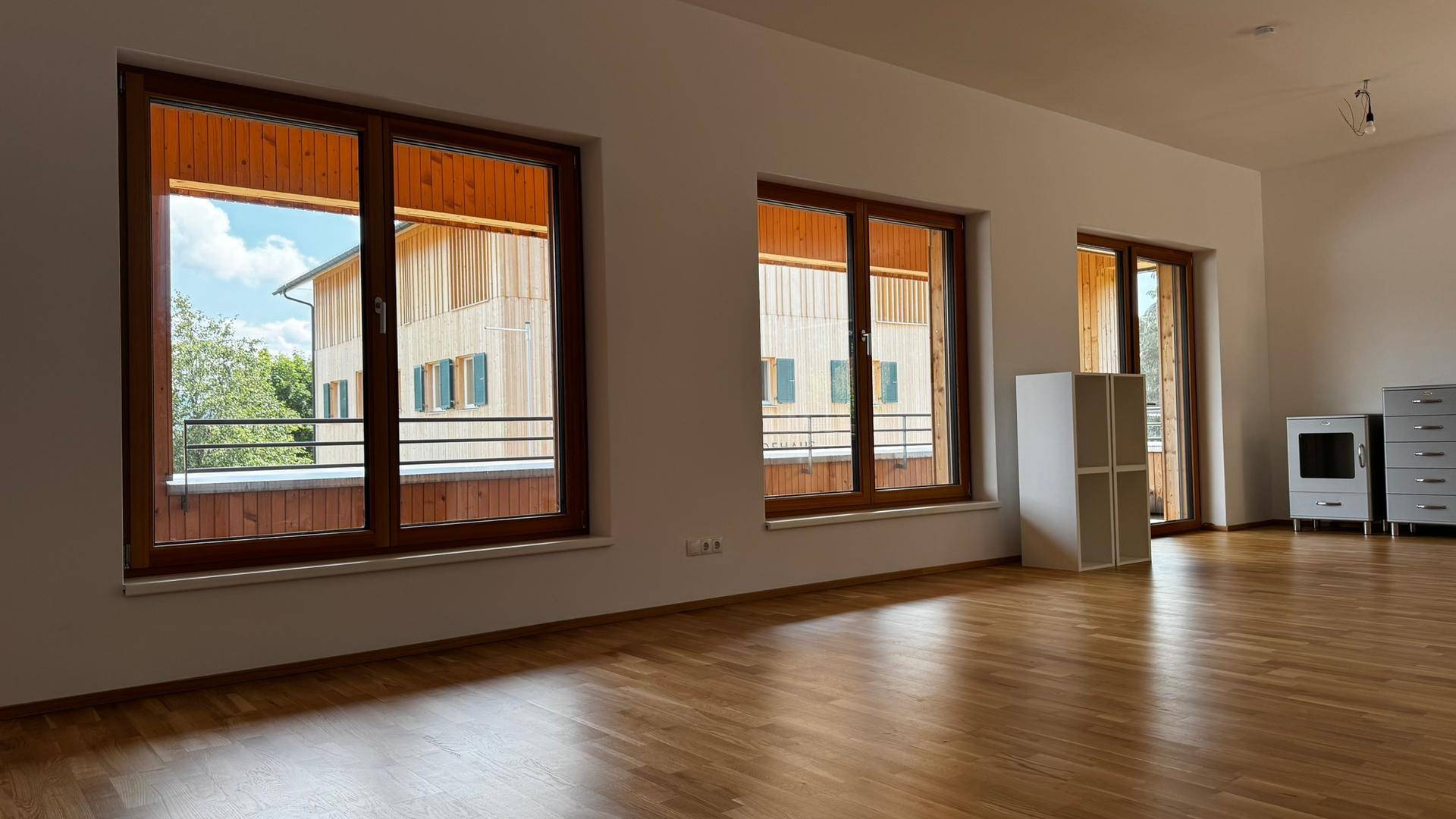 Heller, leerer Wohnraum mit Holzboden und großen Fenstern, die den Blick auf ein Nachbargebäude freigeben.