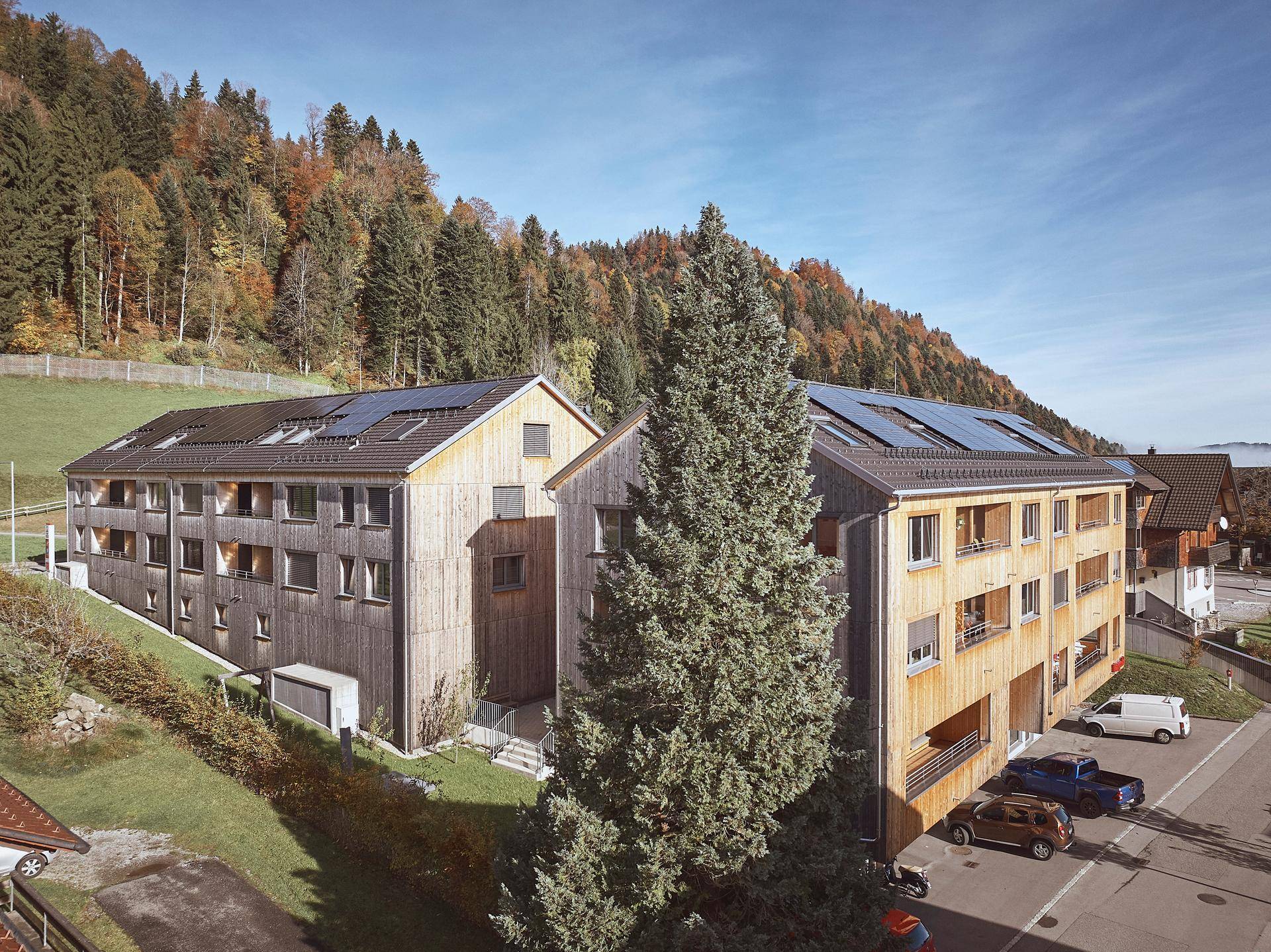 Zwei moderne Gebäude mit Holzfassaden und Solarpaneelen auf den Dächern, umgeben von grüner Natur.