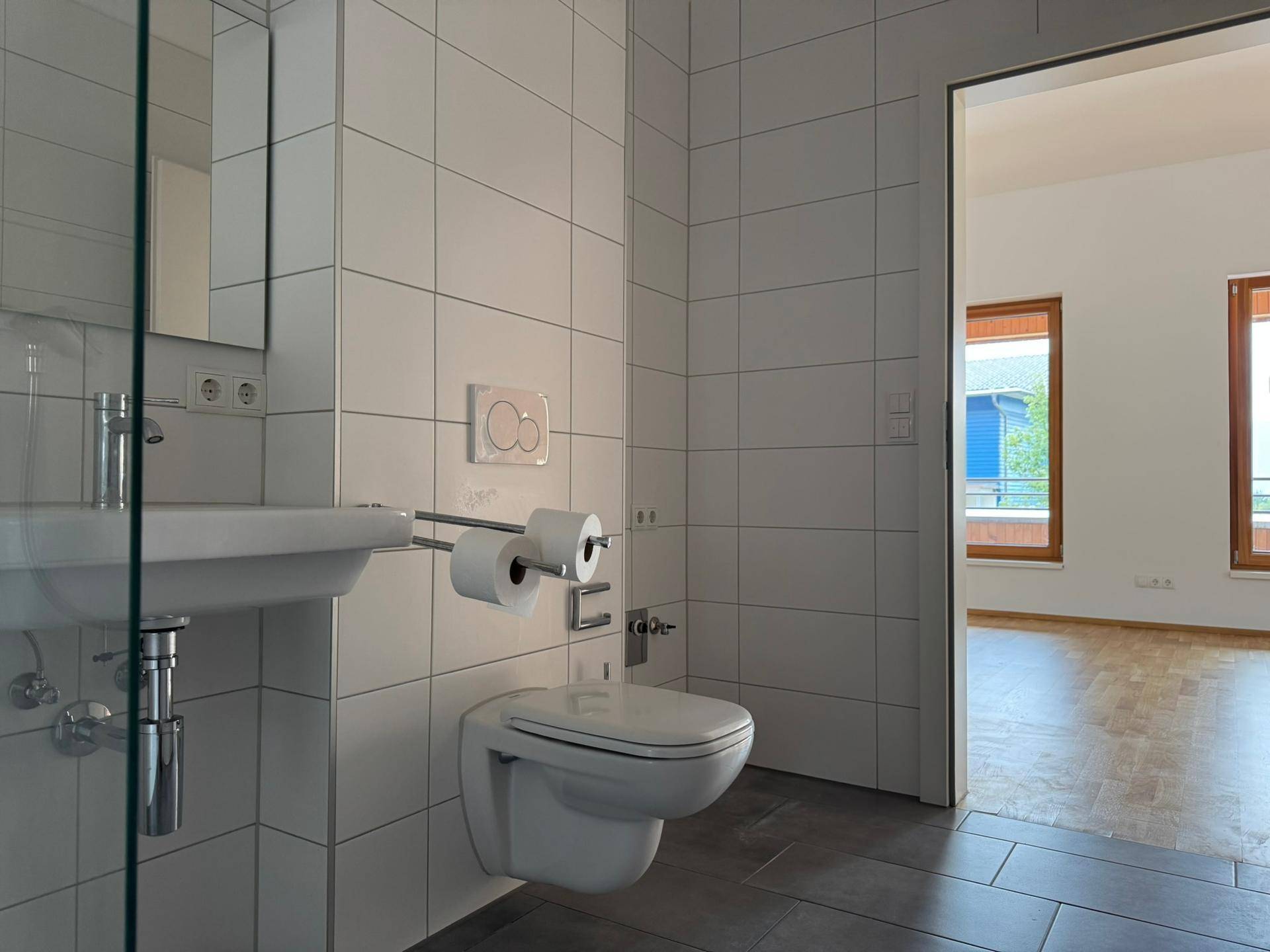 Modernes Badezimmer mit weißen Wandfliesen, dunklem Boden und wandhängendem WC, direkt neben dem Wohnbereich.