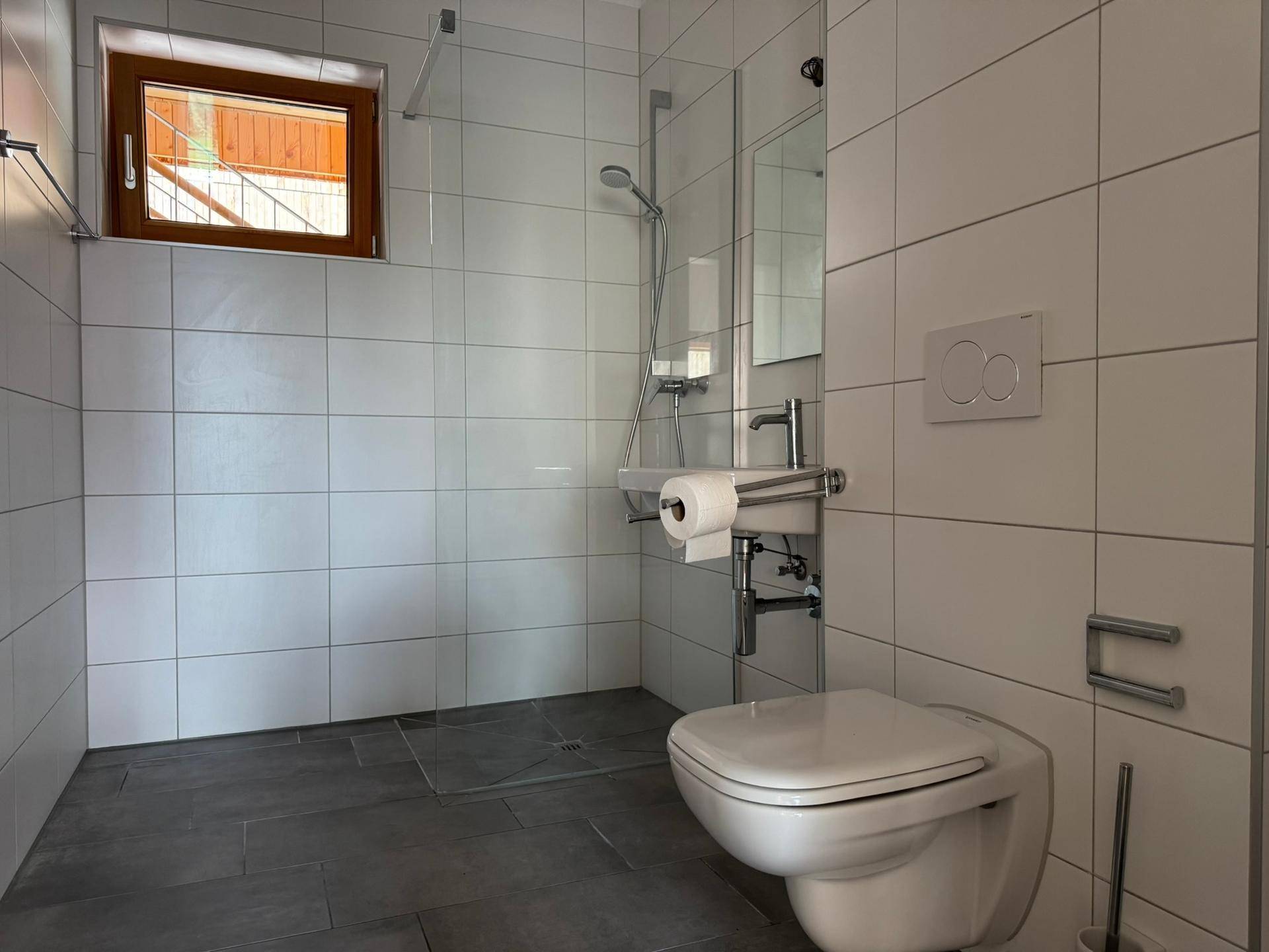 Helles Badezimmer mit ebenerdiger Dusche, wandhängendem WC und einem Fenster für natürliche Belüftung.