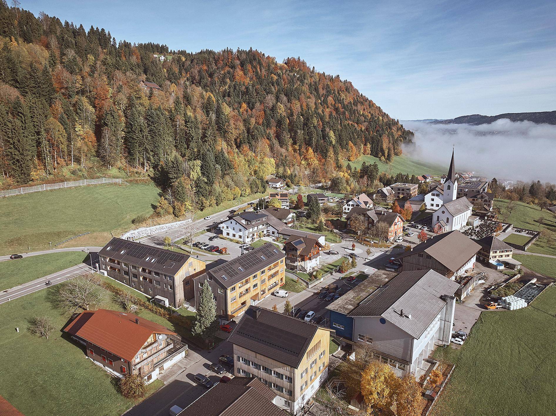 Luftaufnahme eines malerischen Dorfes in einer Herbstlandschaft mit bewaldeten Hügeln und Nebel in der Ferne.