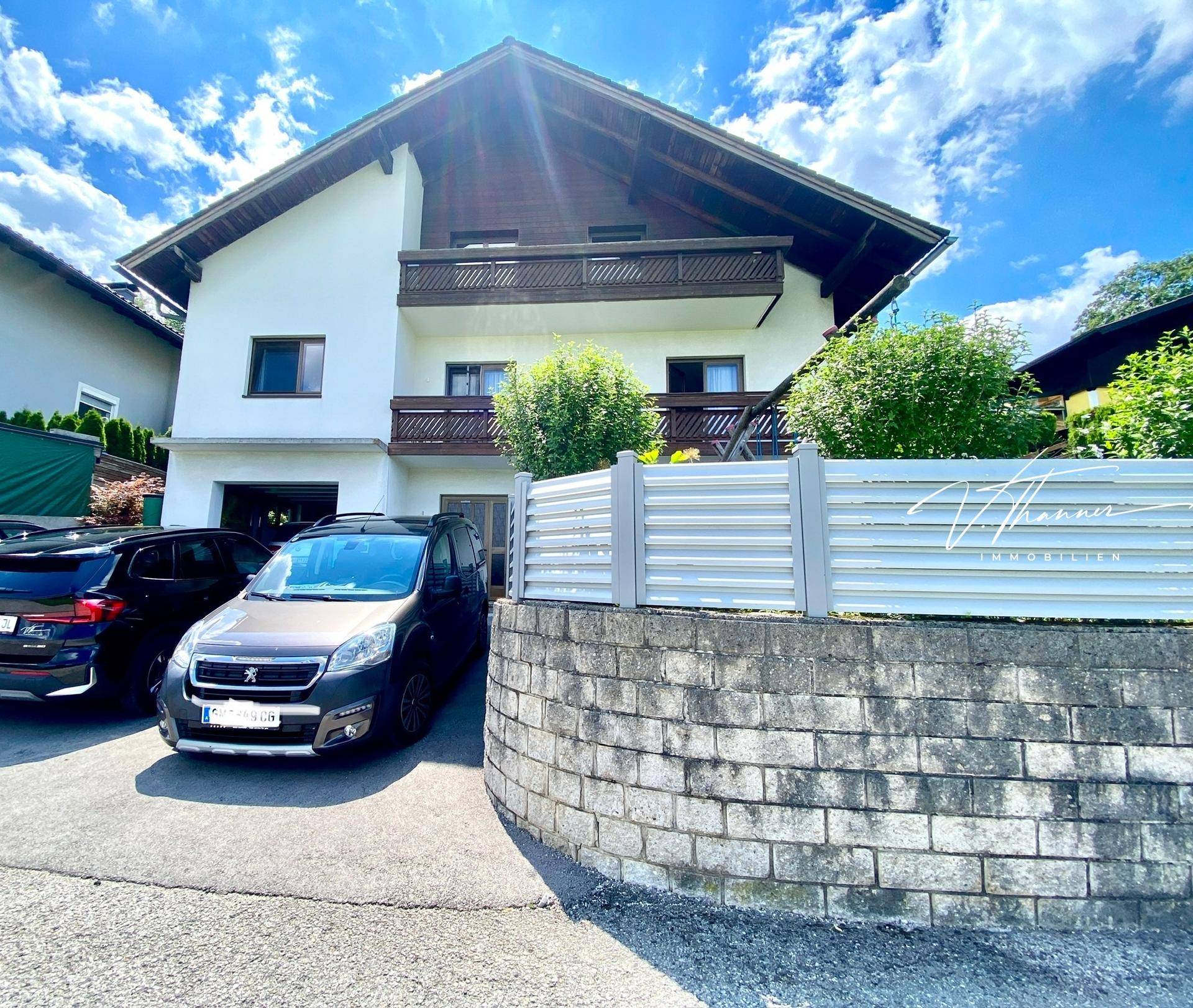 Vorderansicht des Hauses mit Balkonen, Parkplätzen und einer Steinmauer.
