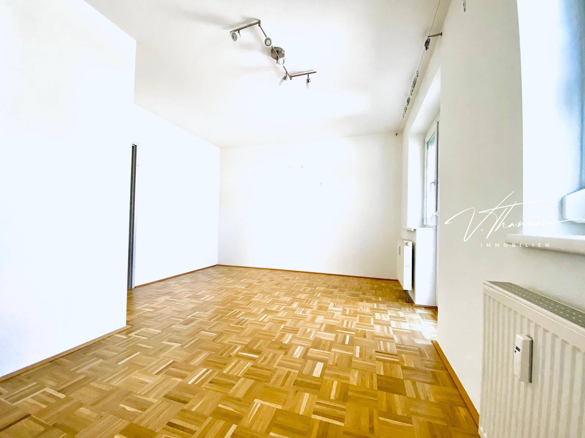 Geräumiges Wohnzimmer mit Parkettboden, weißen Wänden und großen Fenstern für natürliche Beleuchtung.