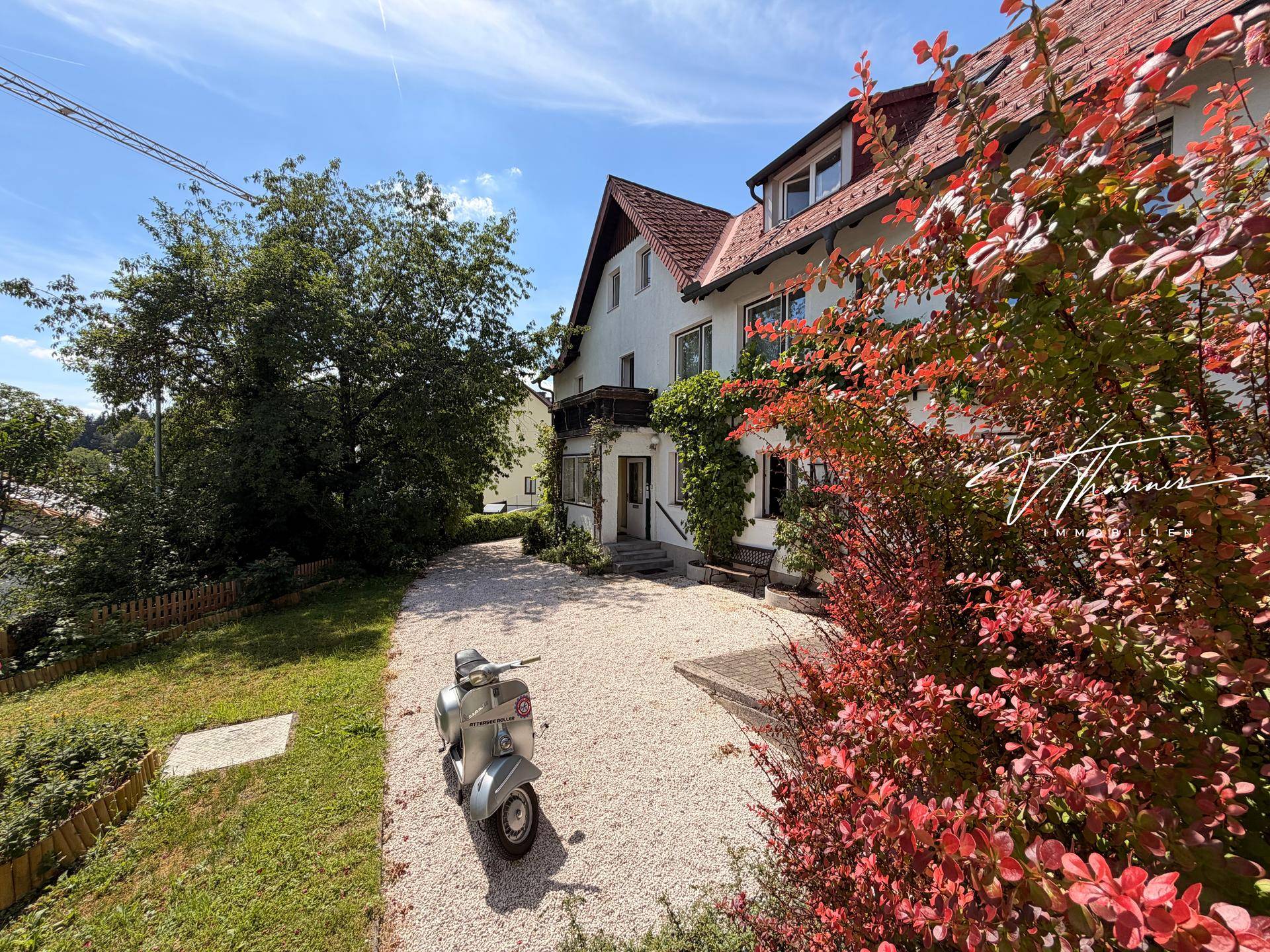 Sonniger Vorgarten mit Kiesauffahrt, blühenden Sträuchern und dem Haupteingang des Hauses.