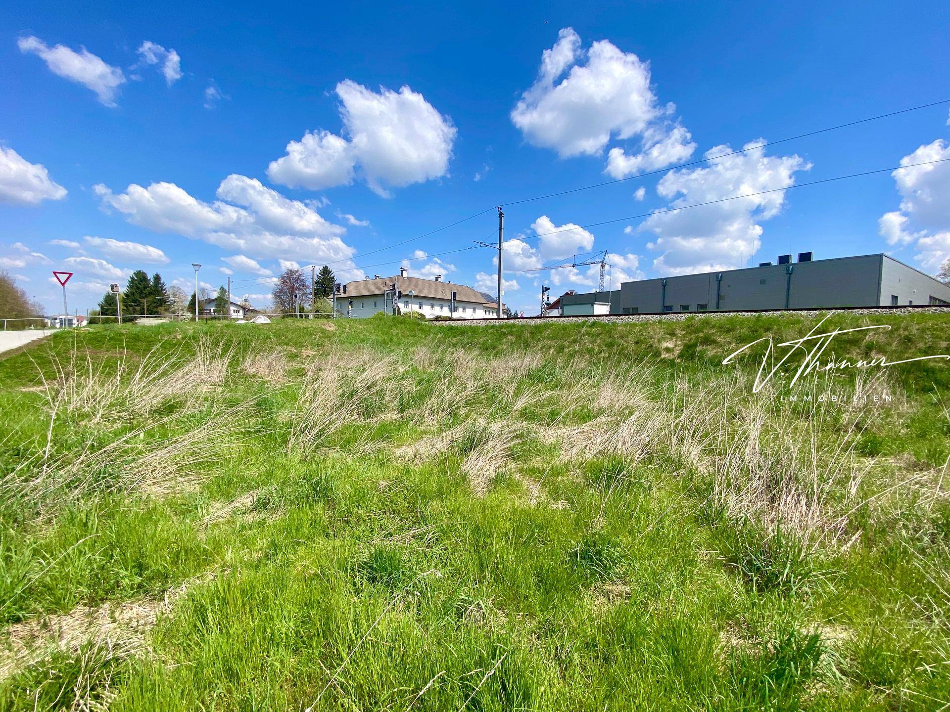 Weitläufige Wiesenfläche mit Gebäuden und Bahngleisen im Hintergrund unter einem strahlend blauen Himmel.