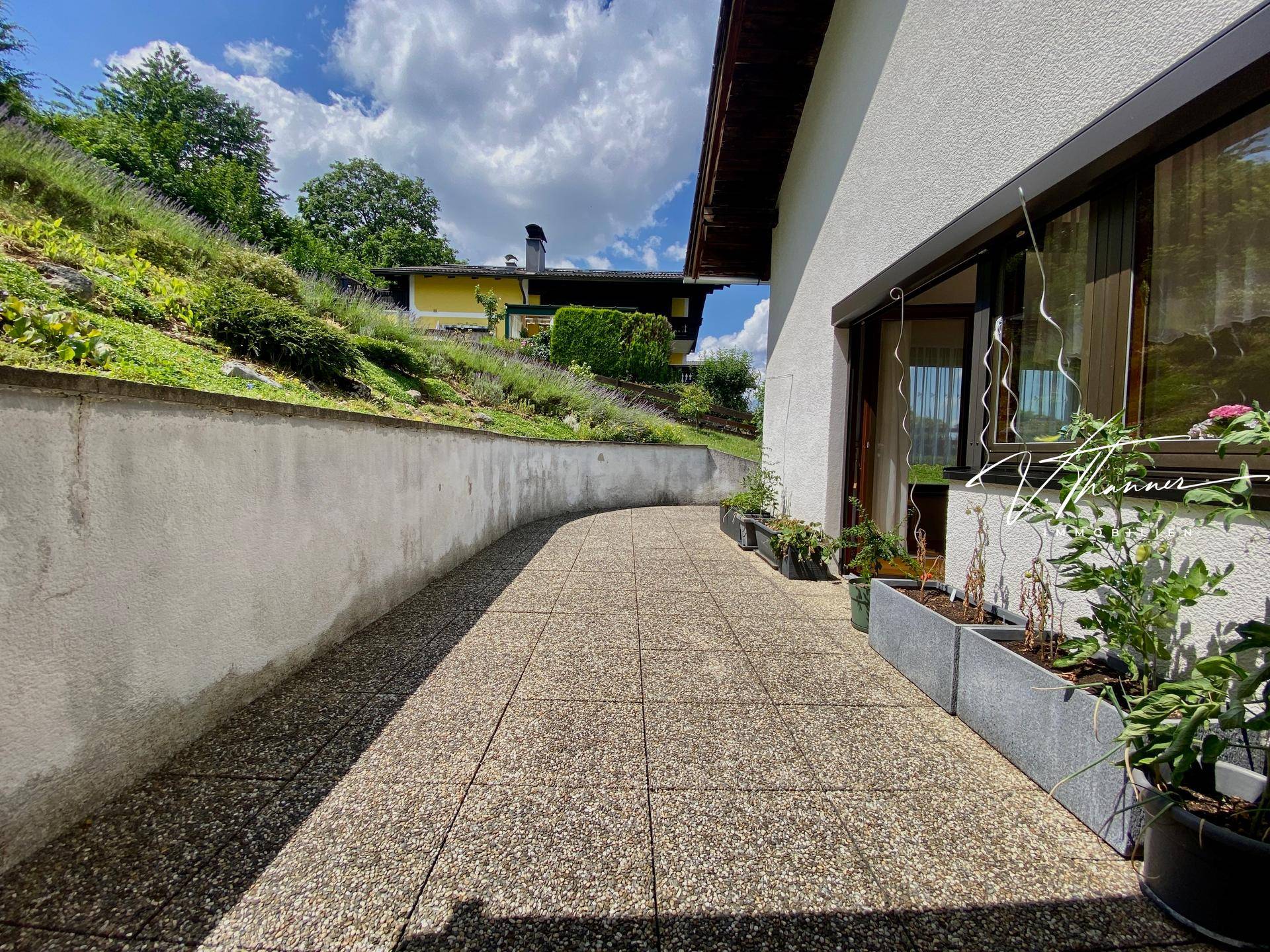 Gepflasterter Terrassenbereich entlang des Hauses mit einer Stützmauer und Pflanzen.