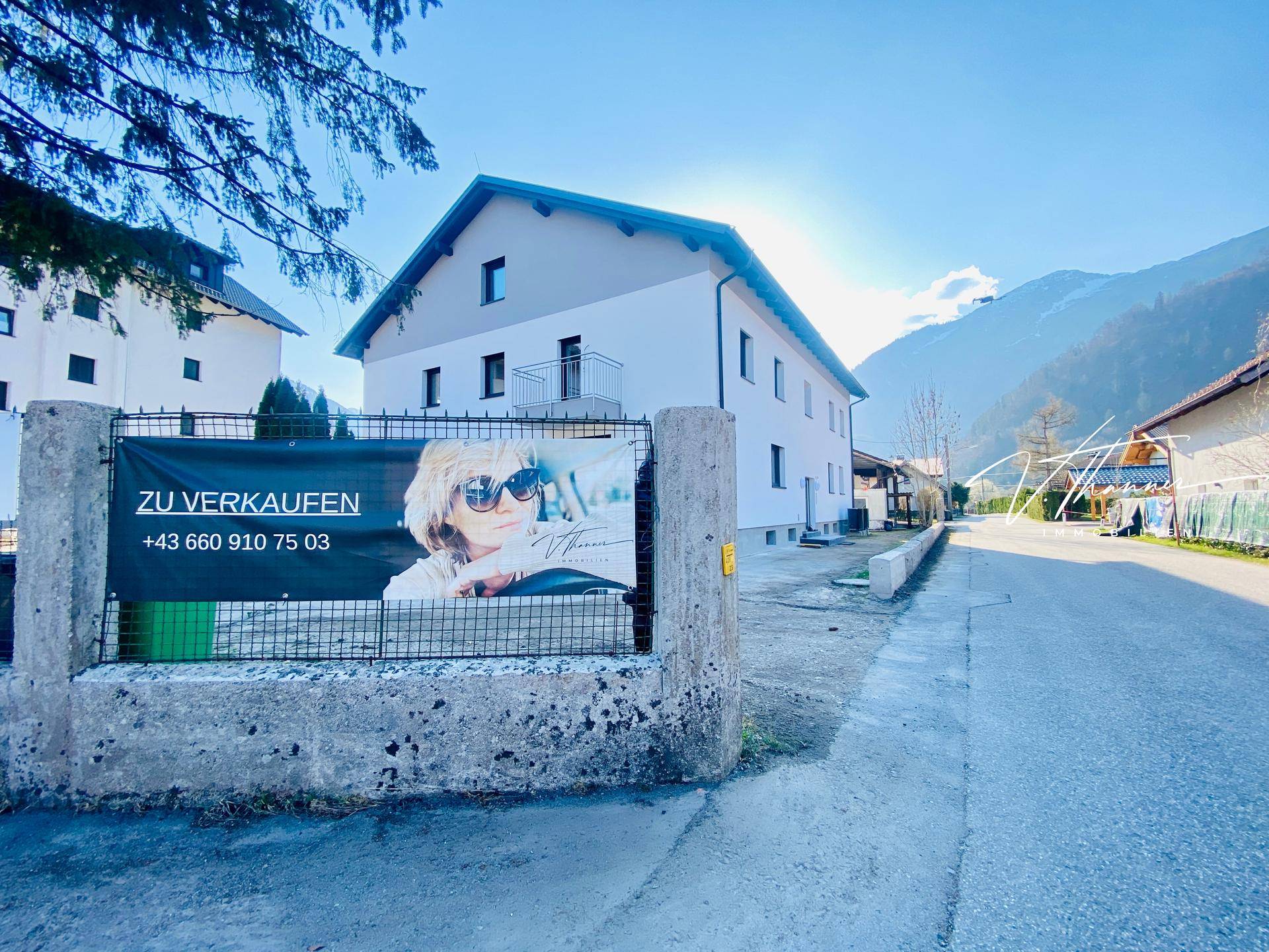 Neubau mit Satteldach und Verkaufsbanner an der Straße, umgeben von Bergen.