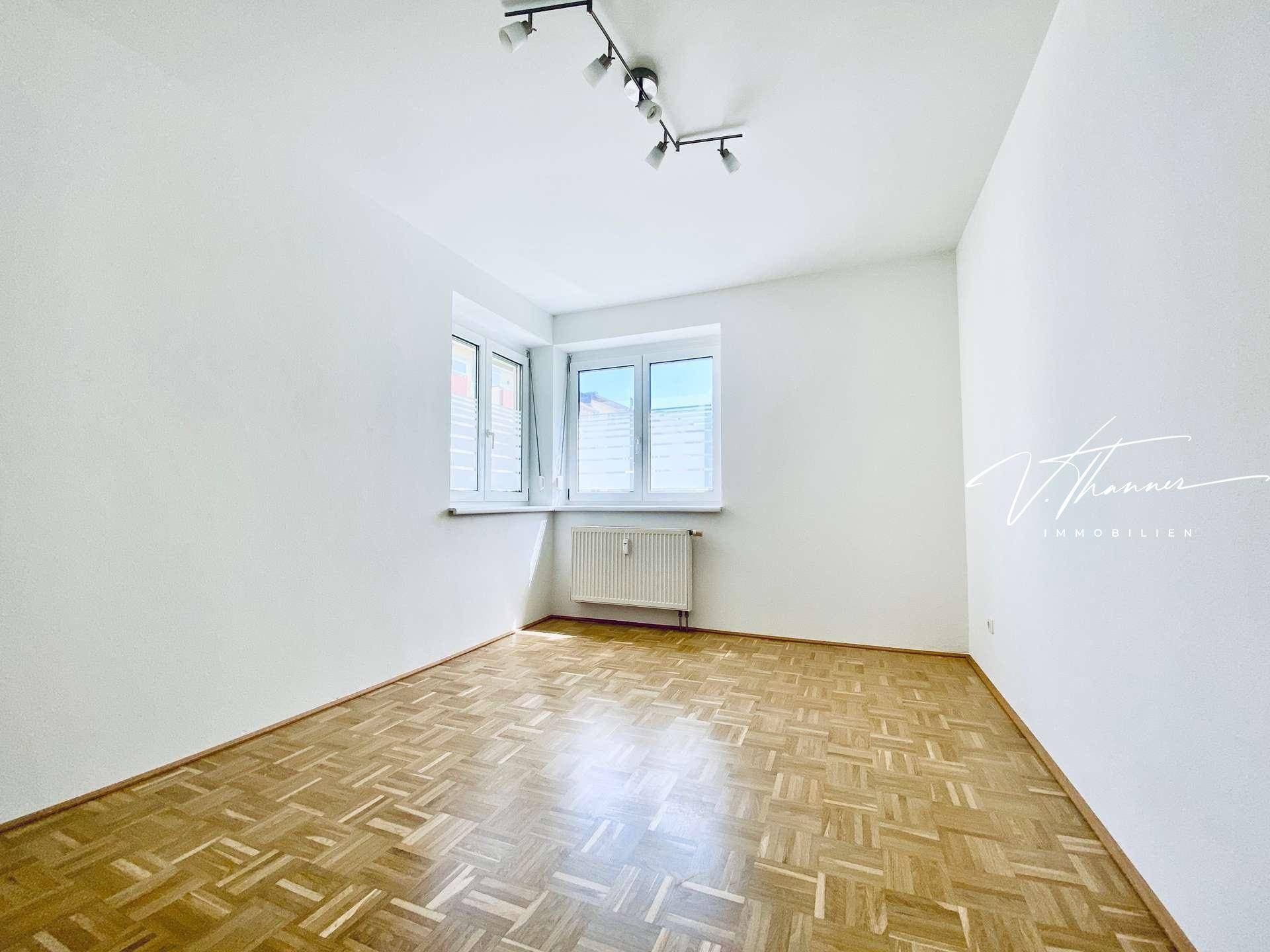 Helles Zimmer mit klassischem Parkettboden, weißen Wänden und zwei Fenstern für viel Tageslicht.