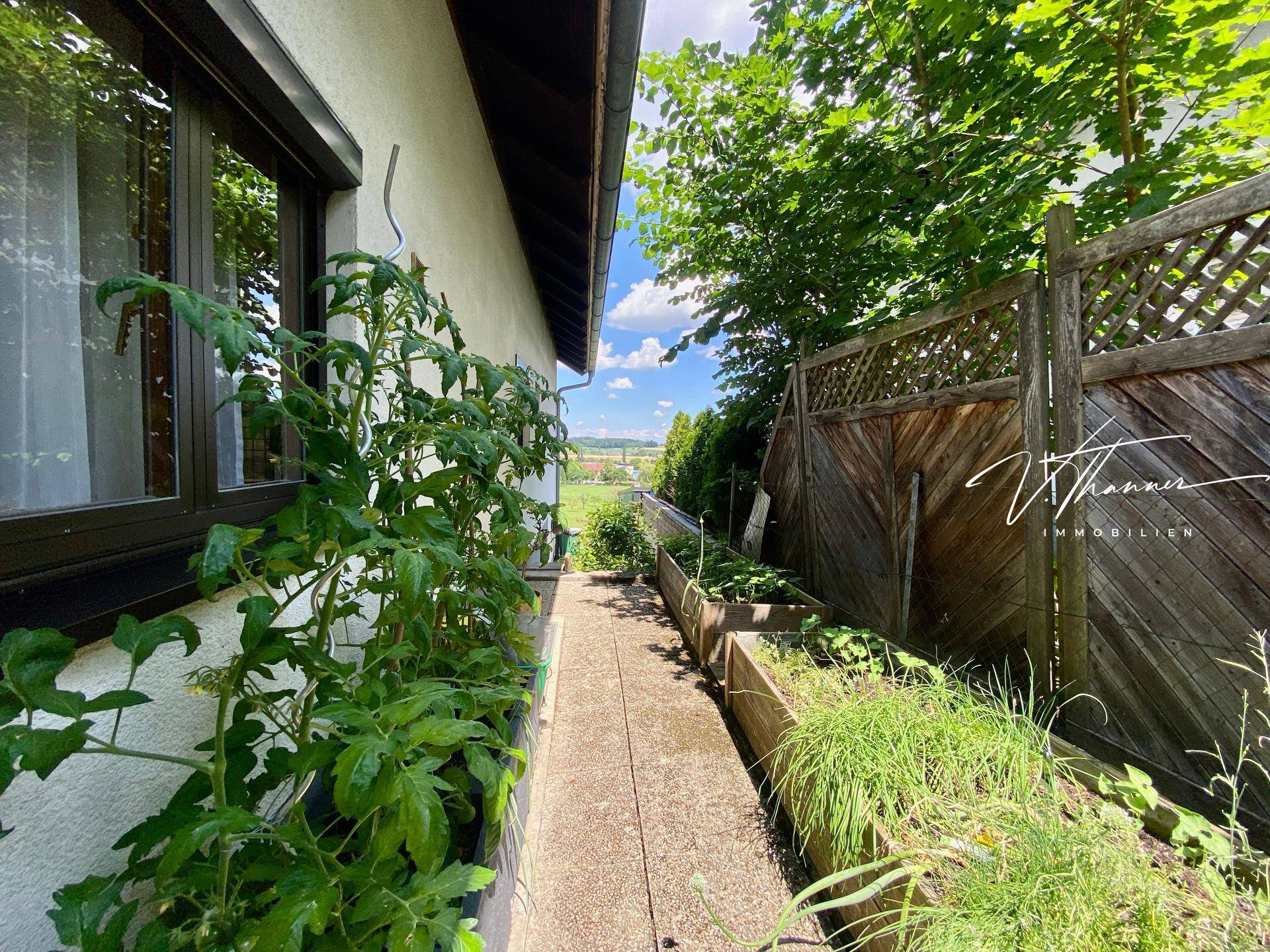Schmaler Gartenweg entlang des Hauses mit Hochbeeten für den Anbau von Pflanzen.