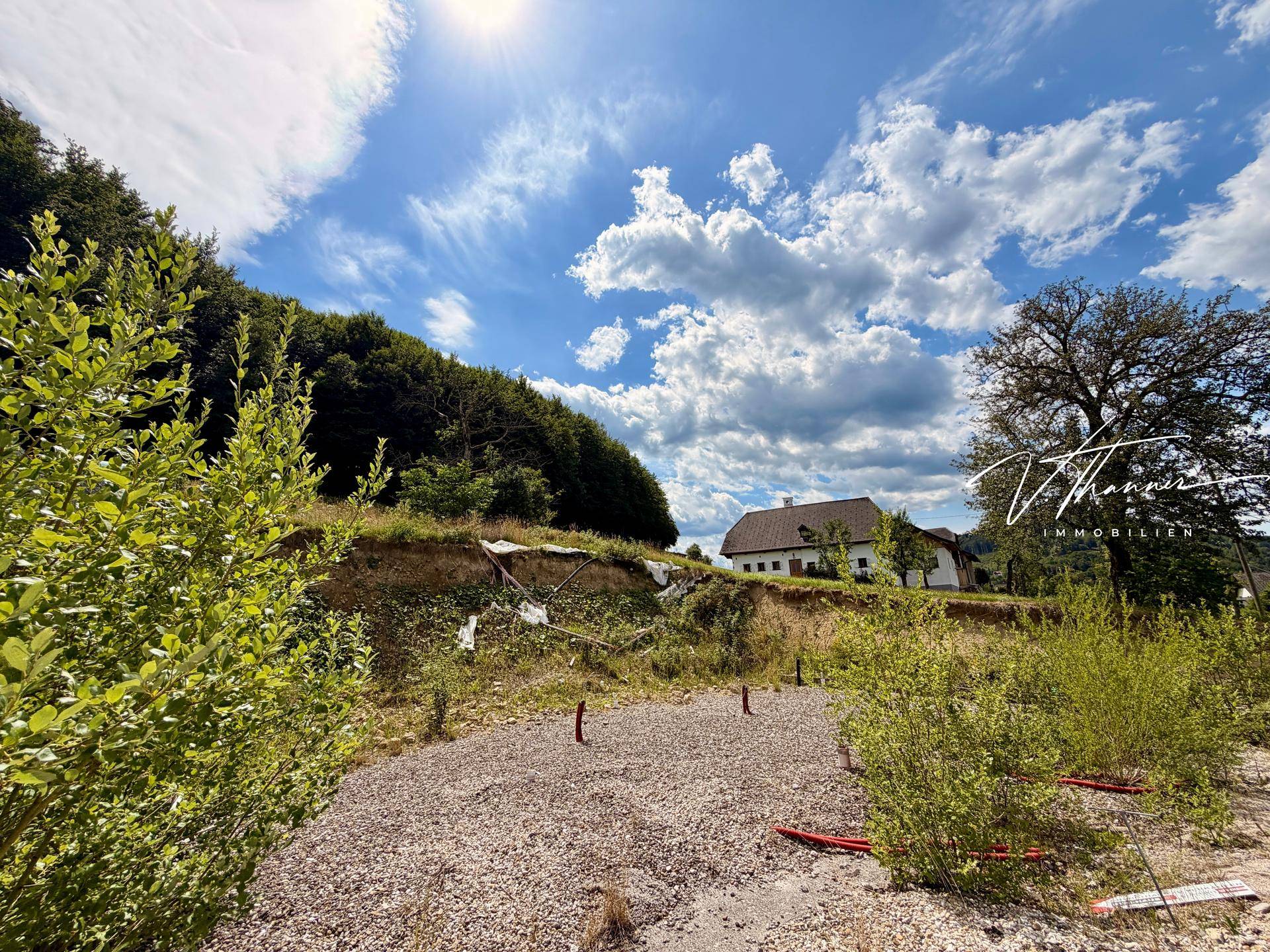 Baustelle mit Schotter und Büschen, umgeben von Wald und einem Wohnhaus im Hintergrund.