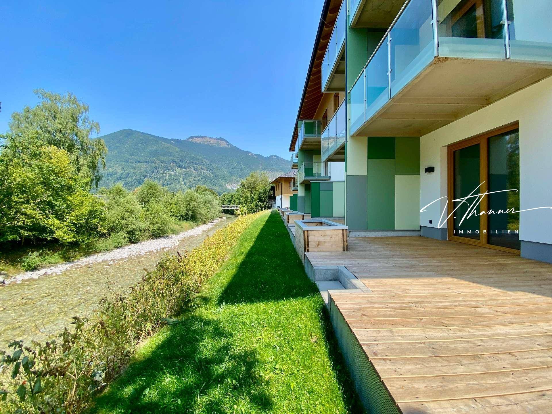 Lange Holzterrasse entlang des Flusses mit Blick auf die Berge und die grüne Landschaft.
