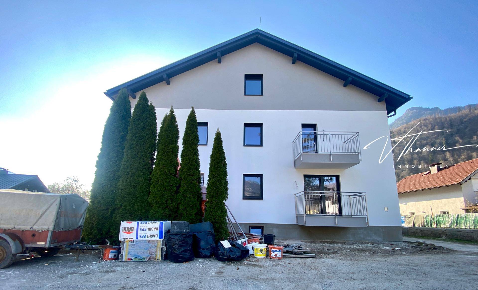 Seitenansicht des Neubaus mit Balkonen und hohen Bäumen, umgeben von Baustellenmaterial.