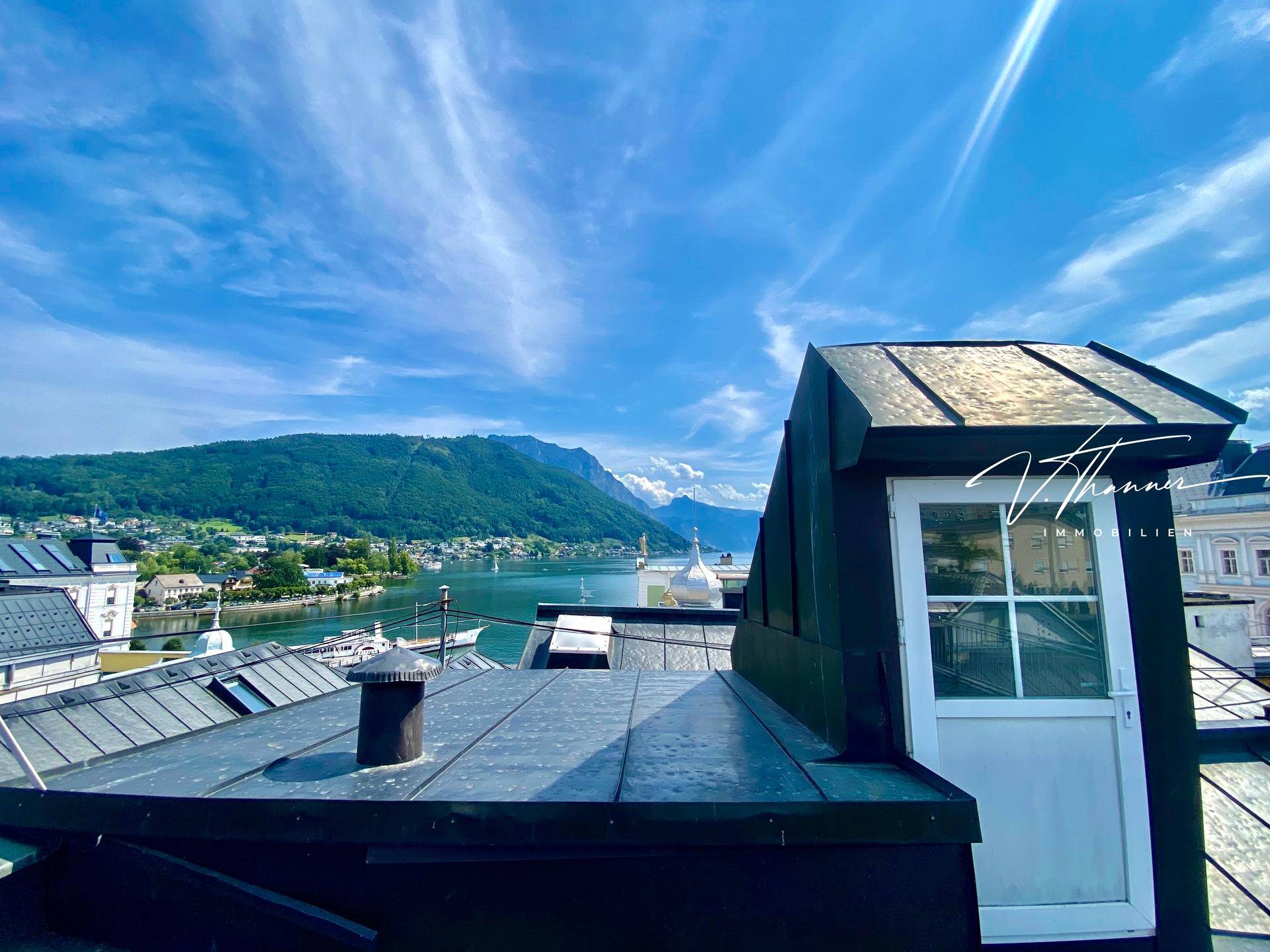 Weitläufiger Blick vom Dach über den See und die umliegenden Berge unter blauem Himmel.