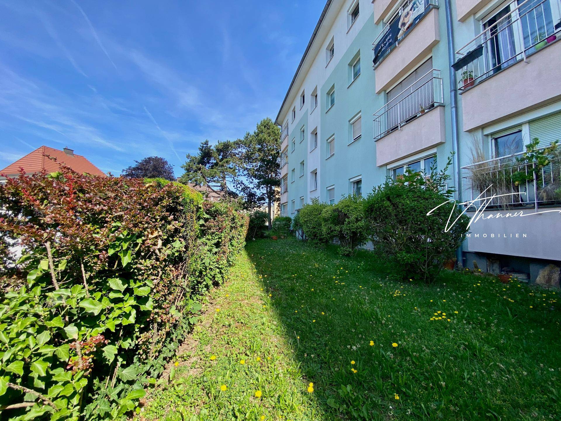 Mehrfamilienhaus mit hellblauer Fassade, Balkonen und gepflegtem Rasen mit Hecke im Vordergrund.