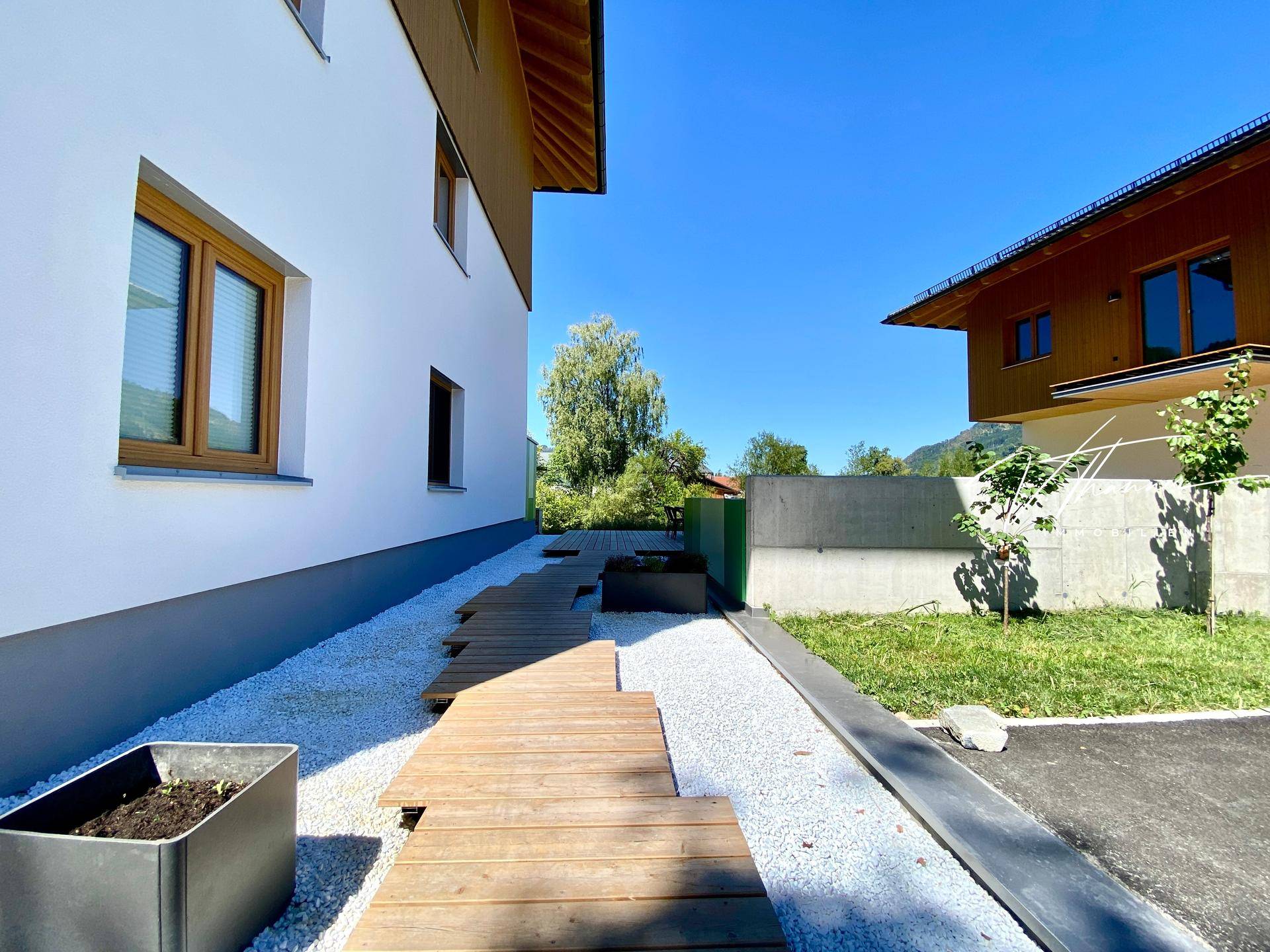 Moderner Gartenweg aus Holzbohlen und weißen Steinen zwischen zwei Gebäuden.