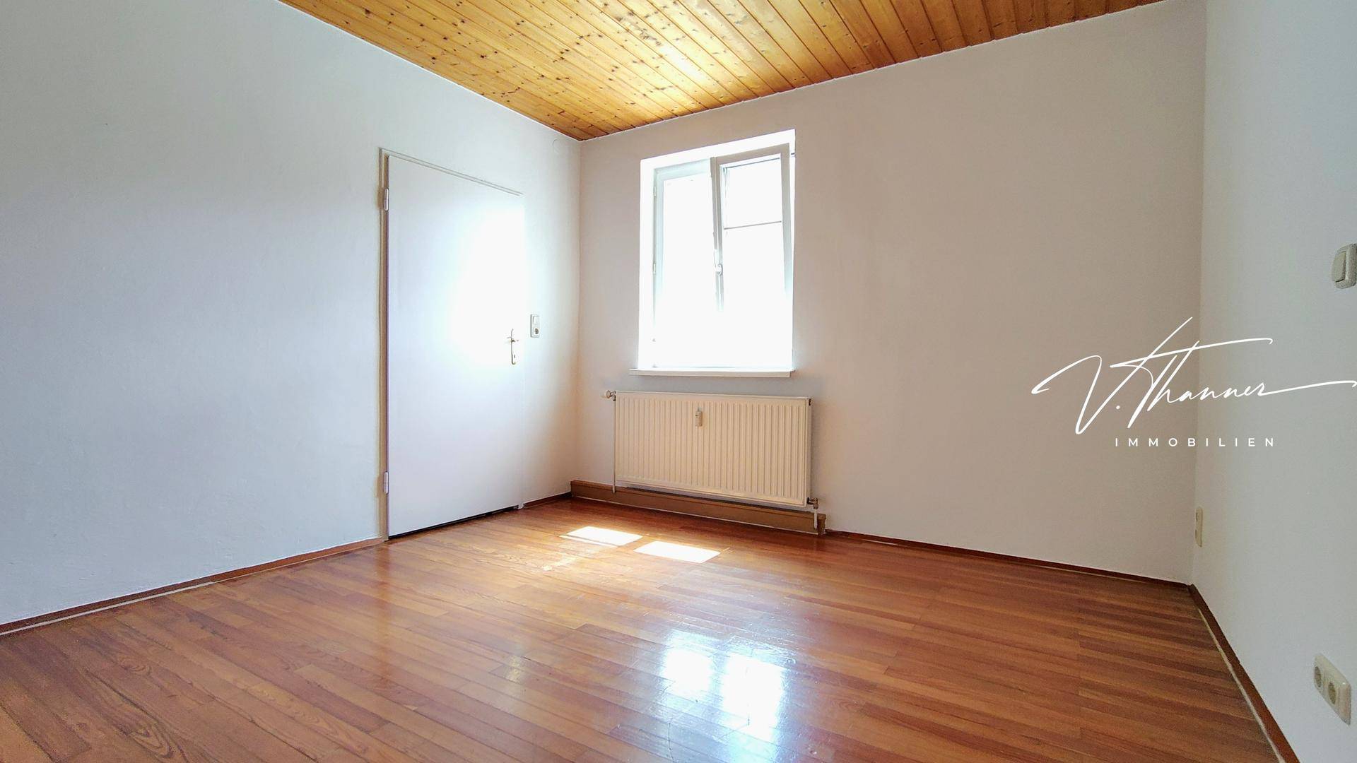 Leeres Zimmer mit dunklem Hartholzboden, Holzdecke und einem Fenster für natürliches Licht.