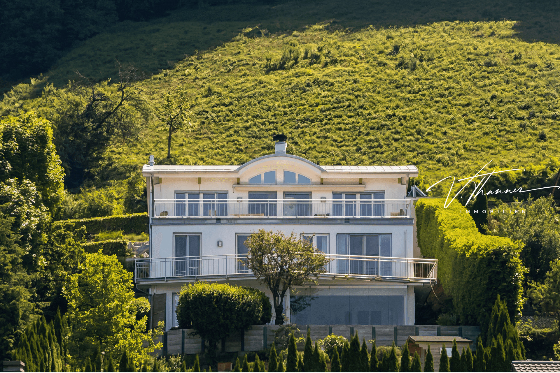 Elegante weiße Villa mit moderner Architektur und mehreren Balkonen, eingebettet in eine grüne Hanglage.
