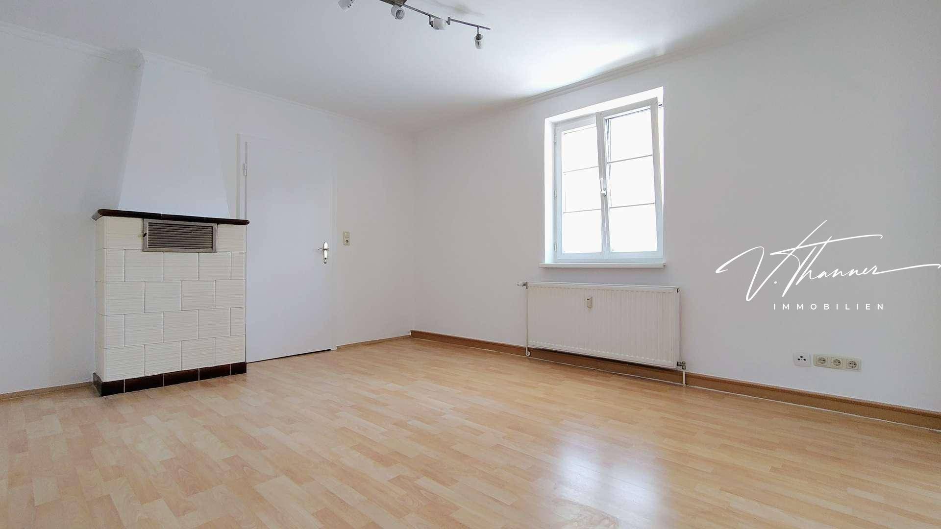 Leeres Zimmer mit hellem Laminatboden, Fenster und einem traditionellen Kachelofen.