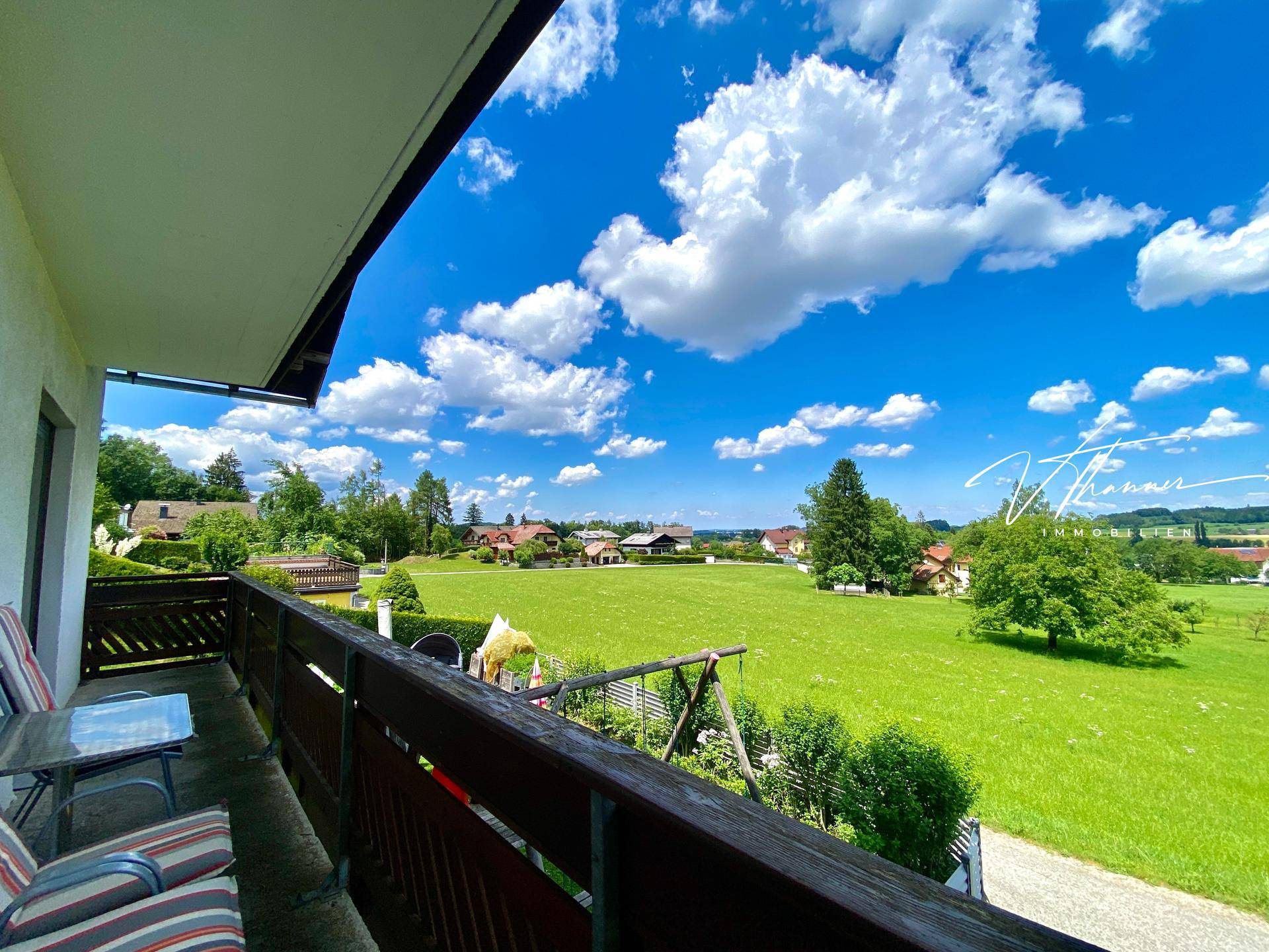 Balkon mit Sitzgelegenheiten und weitem Blick über grüne Felder und die ländliche Umgebung unter blauem Himmel.