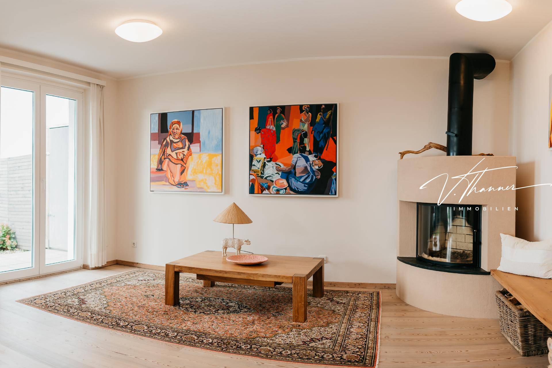 Wohnzimmer mit Kamin, Holztisch und farbenfrohen Kunstwerken, Zugang zum Außenbereich.