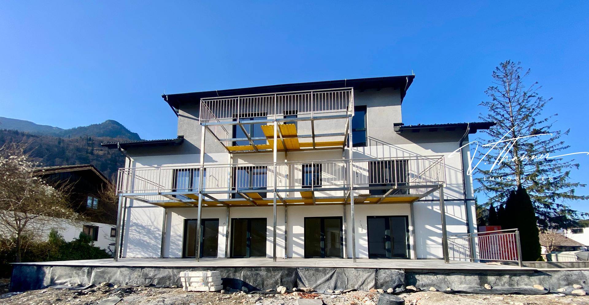 Neubau mit mehreren Balkonen und Gerüst, umgeben von Bergen unter blauem Himmel.