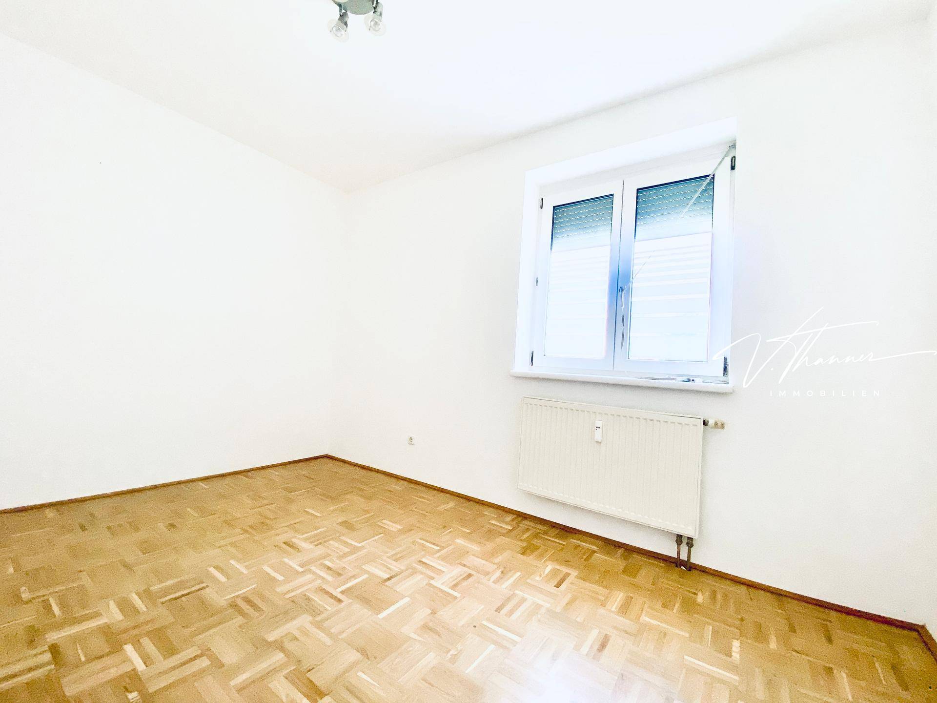 Leeres Zimmer mit Fischgrätparkett, weiß gestrichenen Wänden und einem Fenster mit Rollladen.