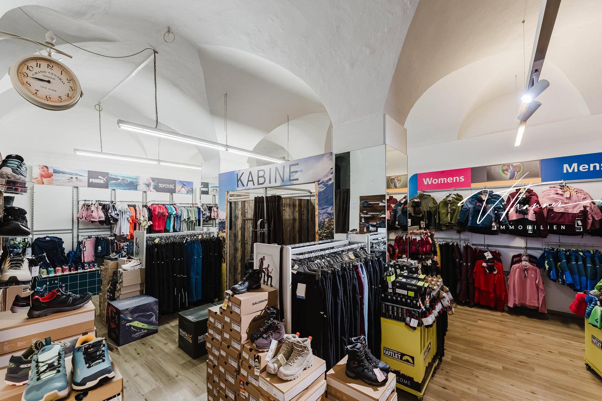 Innenansicht eines Geschäftsraums mit gewölbter Decke und verschiedenen Auslagen für Kleidung und Schuhe.