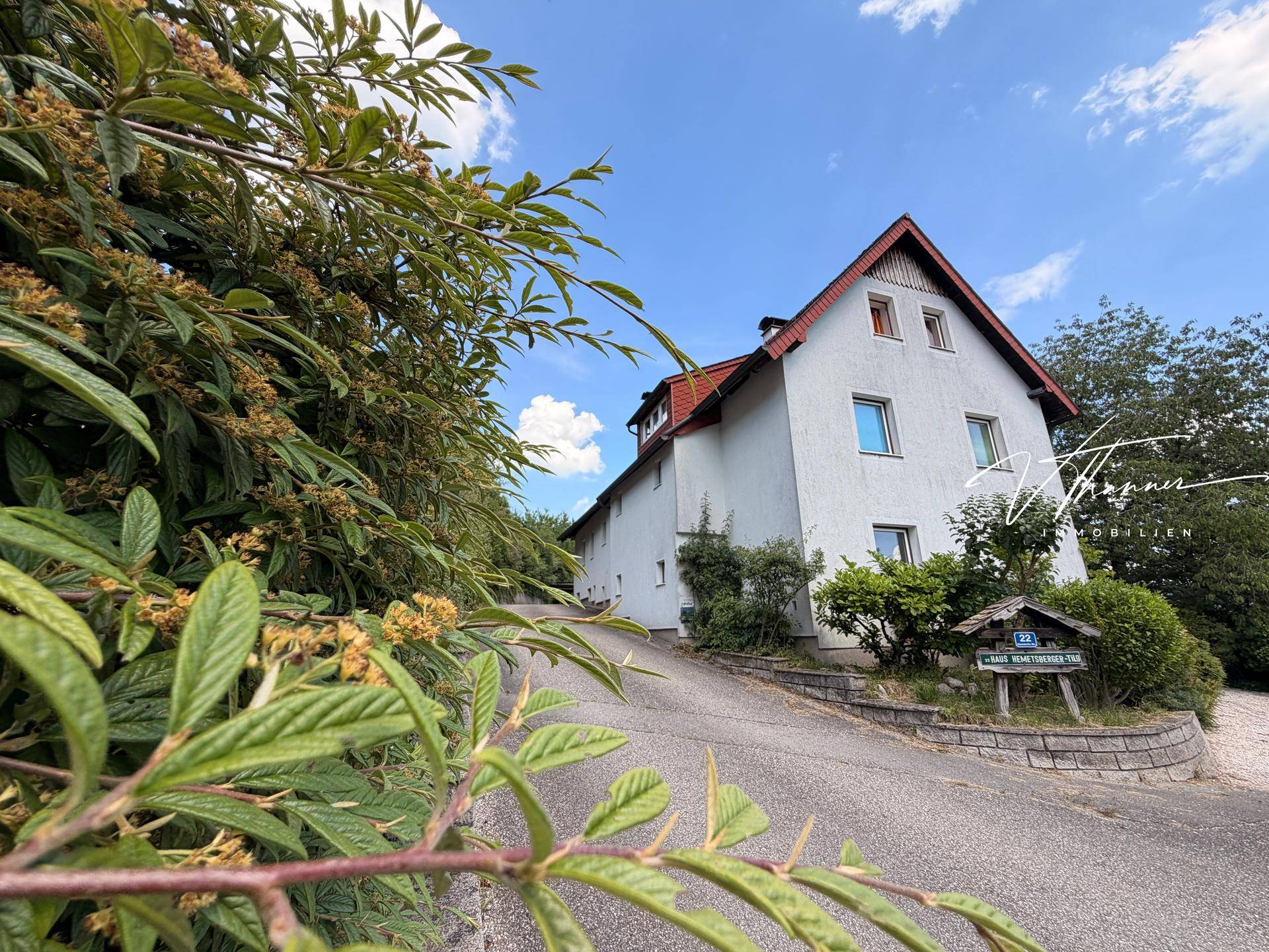 Zufahrt zum Haus mit gepflegter Fassade, Satteldach und umgebender grüner Bepflanzung.