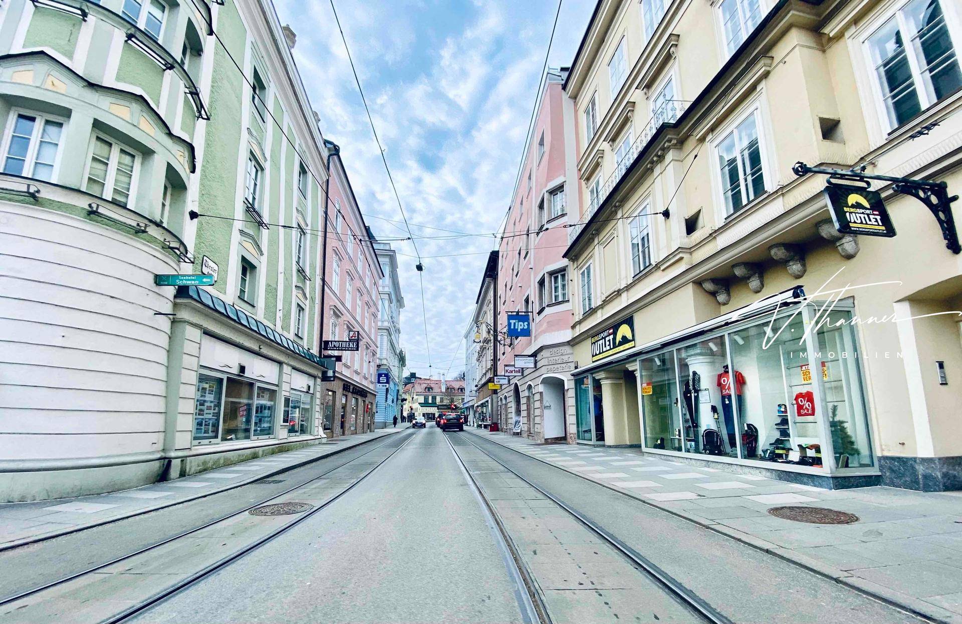 Belebte Straße mit historischen Gebäuden, Geschäften und Straßenbahnschienen unter bewölktem Himmel.