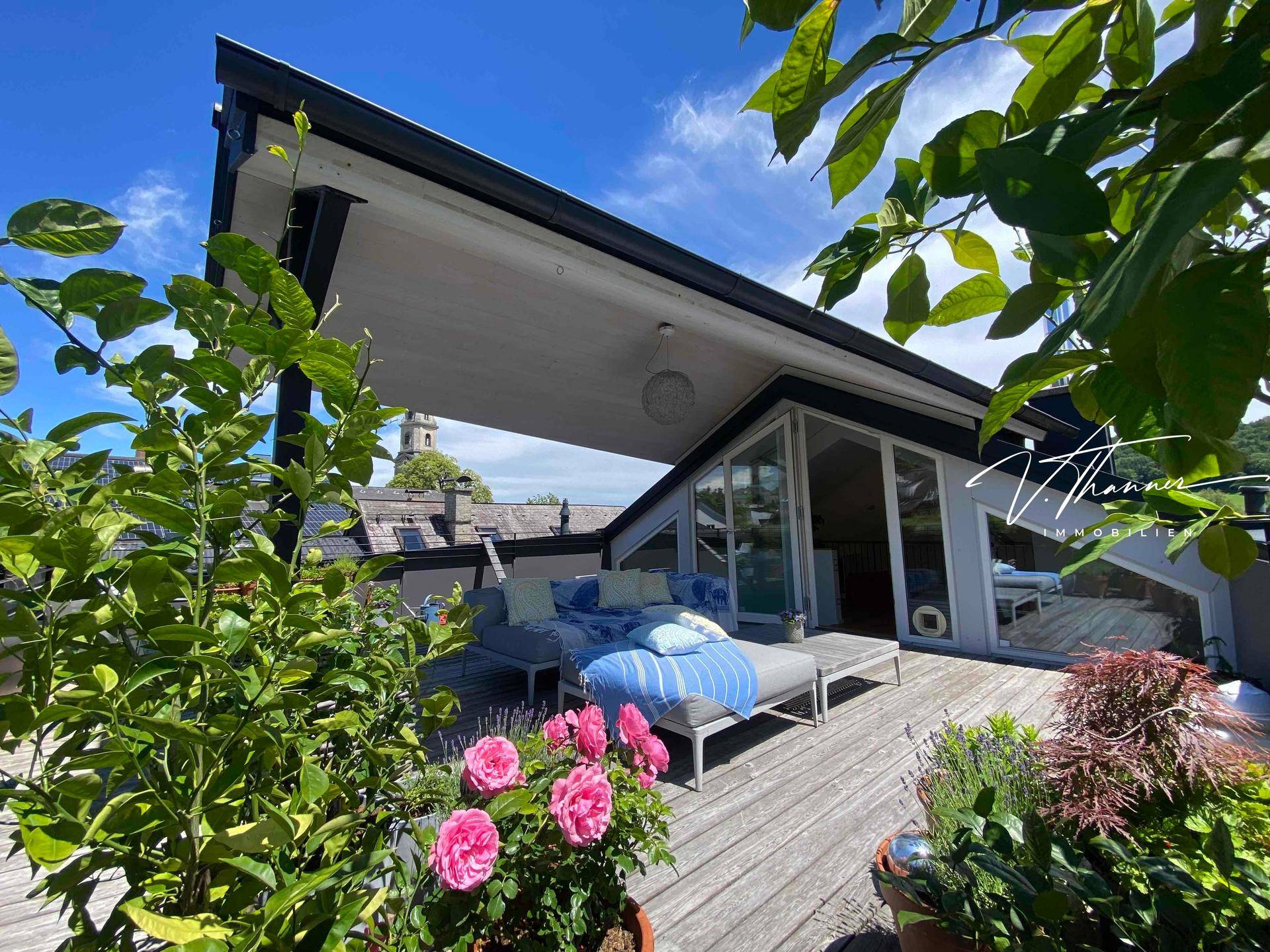 Sonniger Terrassenbereich mit Holzdeck, Pflanzen und Blick auf die Umgebung.