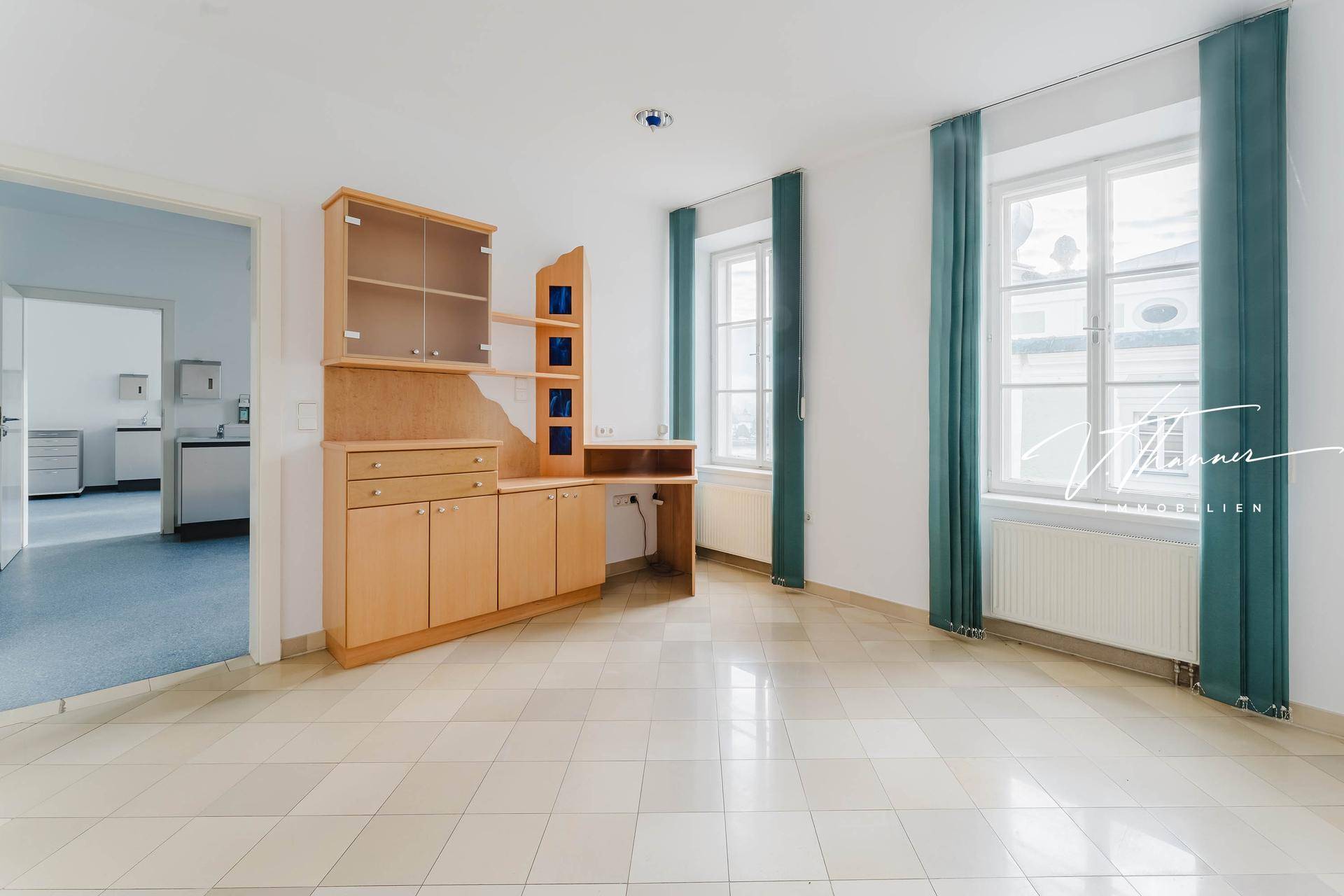 Helles Büro mit Fliesenboden, Holzschrank und großen Fenstern mit Blick nach draußen.