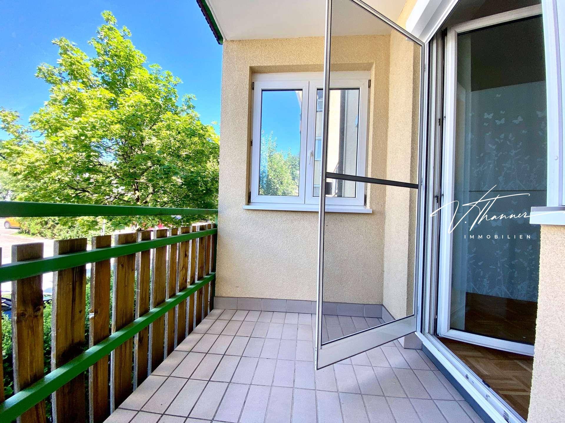 Heller Balkon mit Fliesenboden und Zugang zum Innenraum, umgeben von viel Grün.