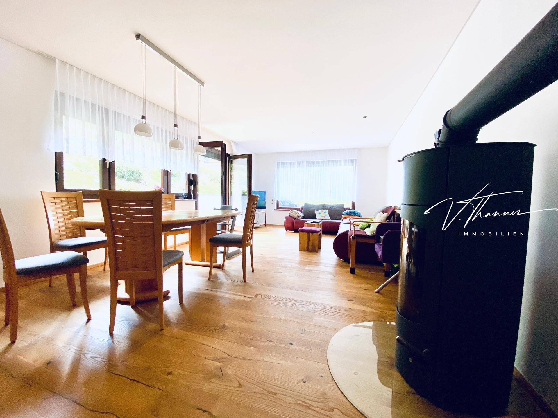 Offener Wohn-Essbereich mit Holzboden, Kaminofen und großen Fenstern zur Terrasse.