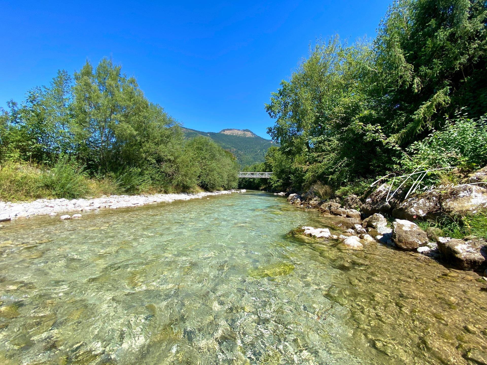 Klarer Flusslauf mit einer Brücke in der Ferne, umgeben von dichtem Grün und Bergen.